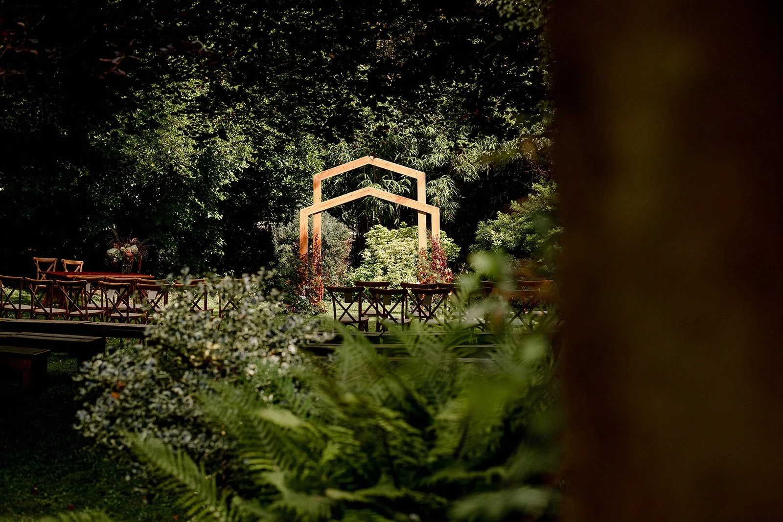 Outdoor wedding ceremony setup in a lush green garden with wooden benches, floral arrangements, and a wooden arch at the center, surrounded by dense foliage.