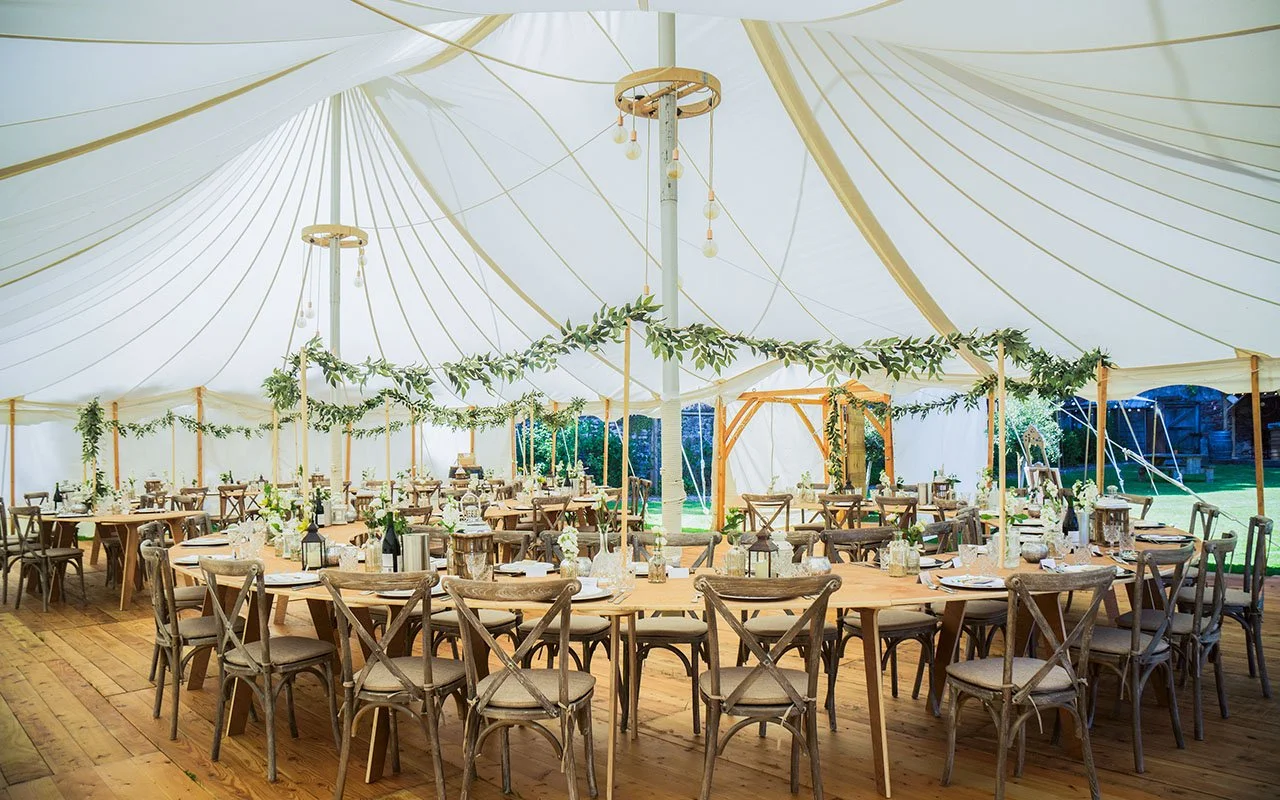 A large, white bespoke marquee decorated with greenery garlands, set up with multiple round tables and wooden chairs for a celebration or wedding reception.