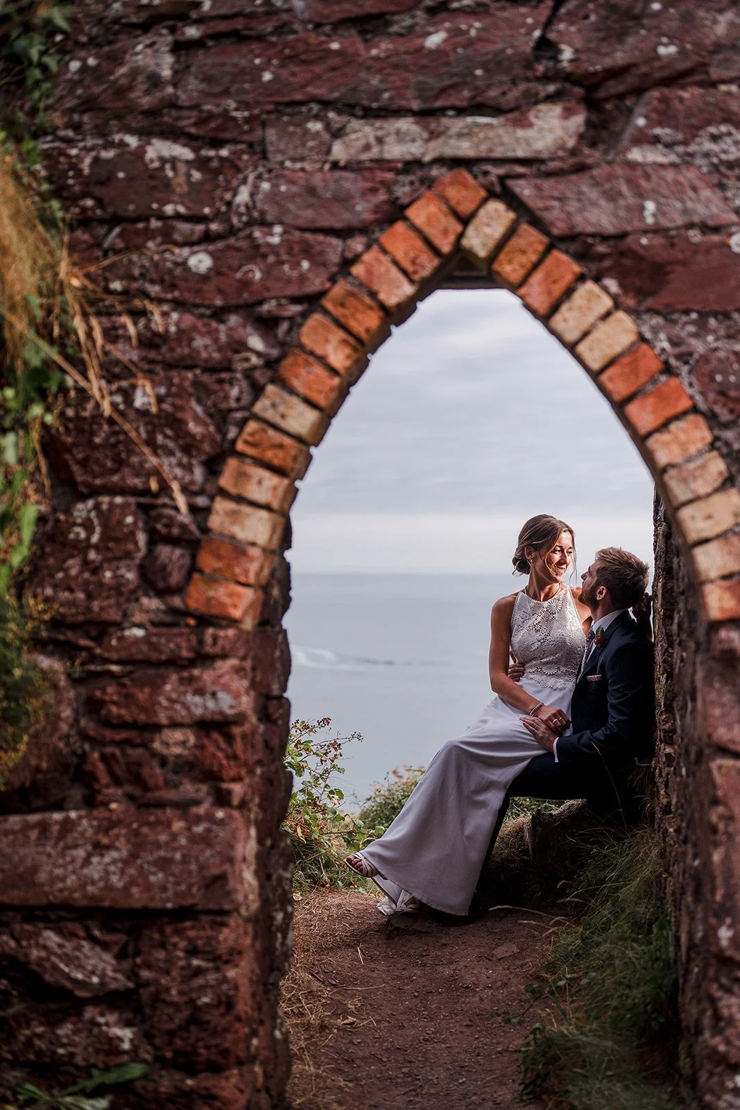 A bride and groom seated together inside a brick archway overlooking the ocean on a cloudy day.