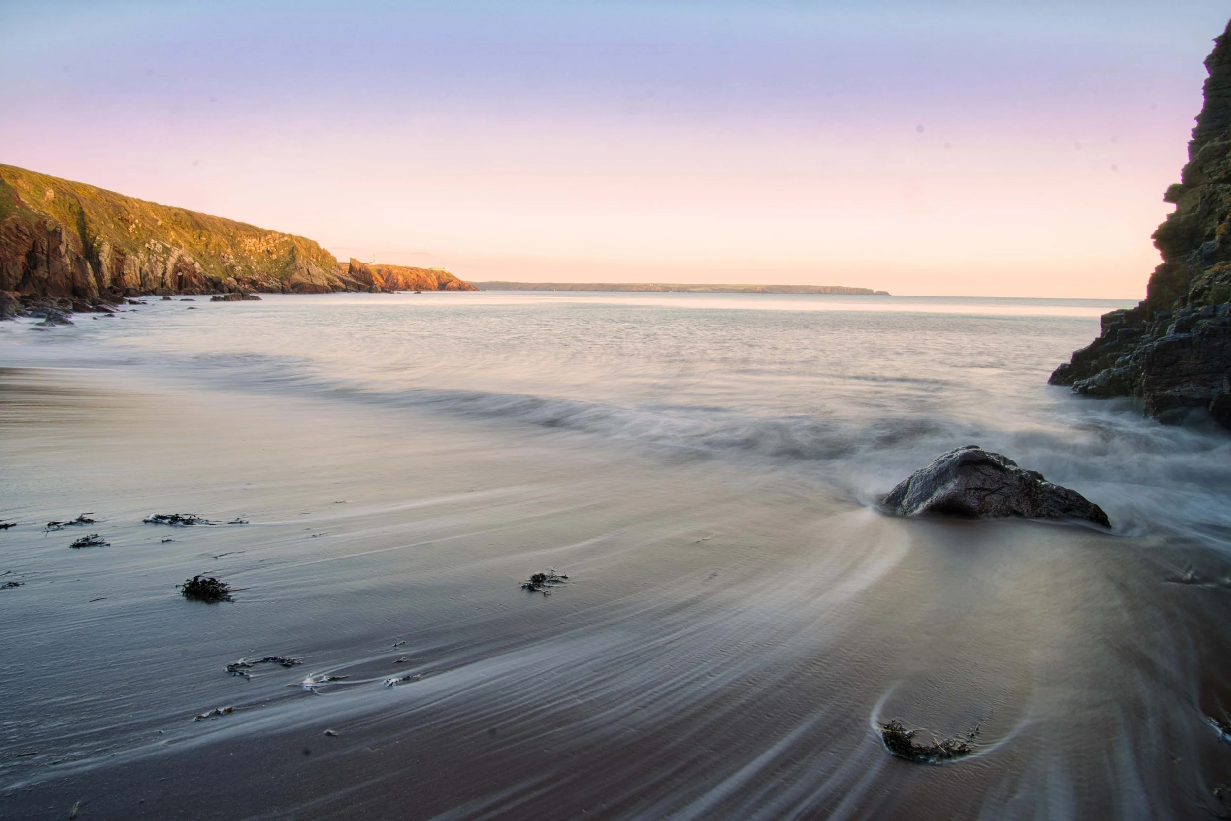 A serene coastal scene at sunset with calm ocean waters, rocky cliffs on either side, and small patches of seaweed on the sandy shore.