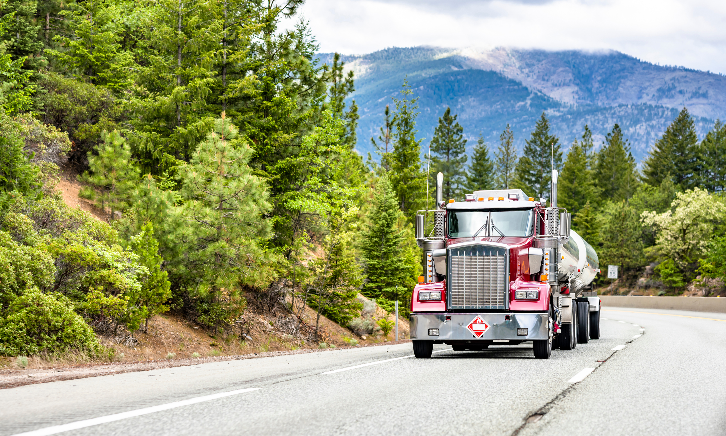 A semi-truck with a red cab and a silver tank drives on a mountain highway surrounded by green pine trees, with mountains in the background under partly cloudy skies.