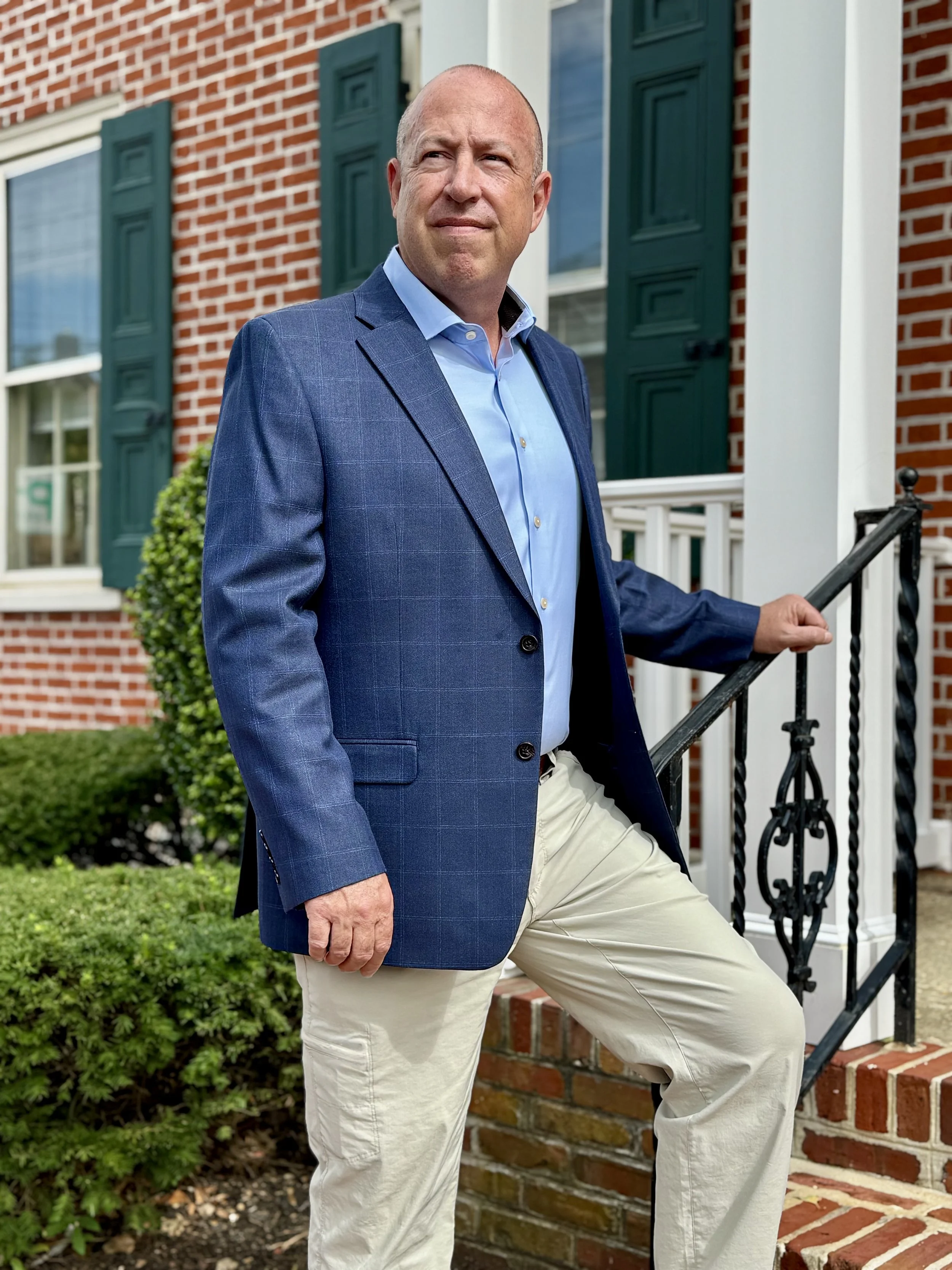 A middle-aged man in a blue blazer and light-colored pants standing on the steps of a brick house with green shutters, surrounded by greenery.