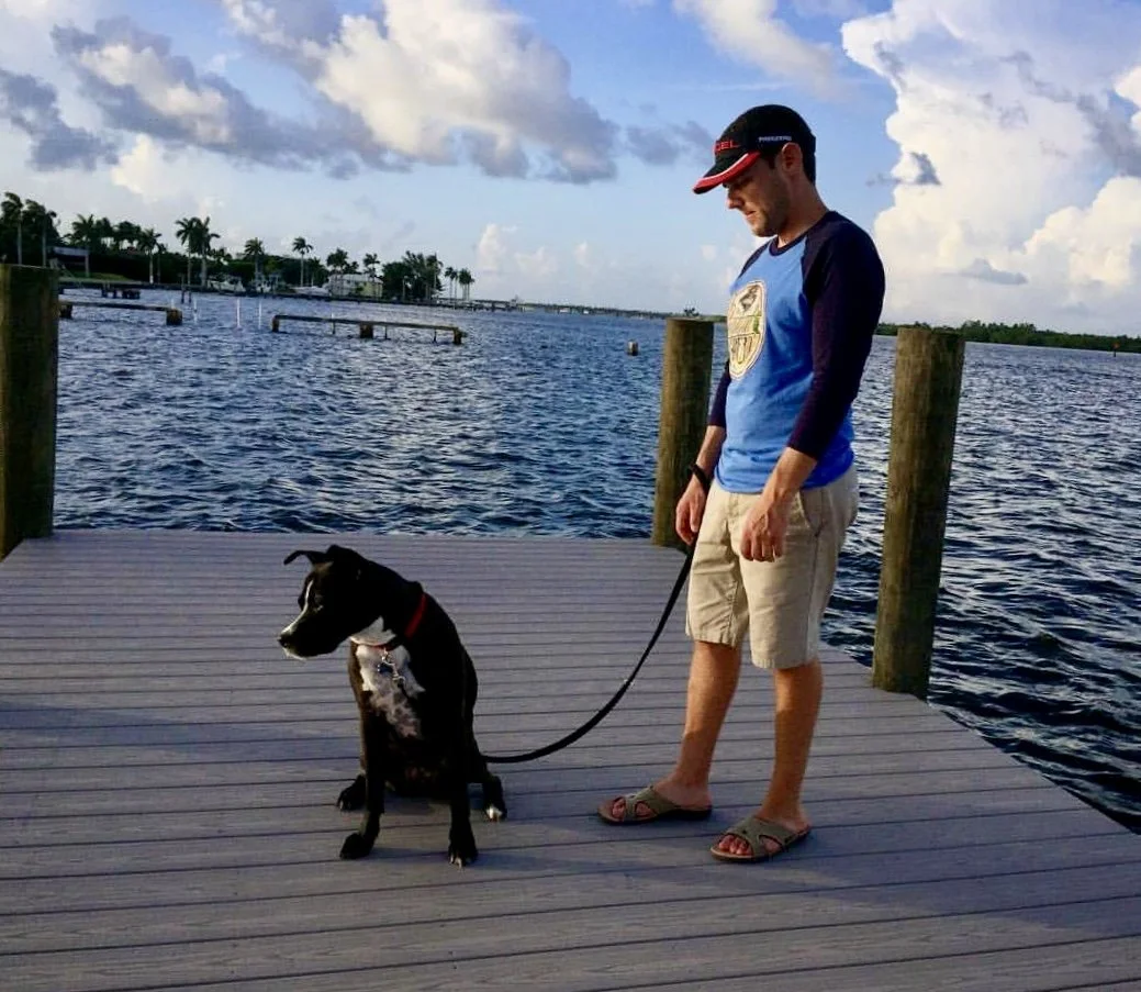 A man and a black dog sit on a wooden dock by the water. The man wears a blue and black long-sleeve shirt, beige shorts, sandals, and a cap. The dog wears a red collar and looks to the side. There are palm trees and houses in the background under partly cloudy skies.