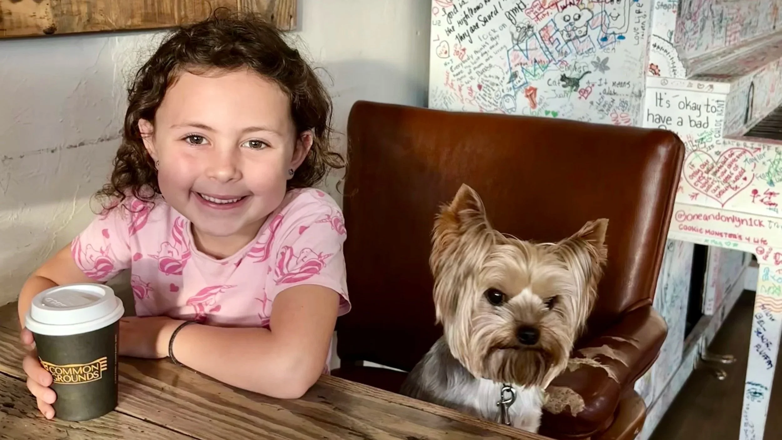 A young girl with curly brown hair wearing a pink shirt with hearts, holding a coffee cup, sitting at a wooden table with a small dog sitting on her right, in a cafe with graffiti-style drawings and text on the wall behind.
