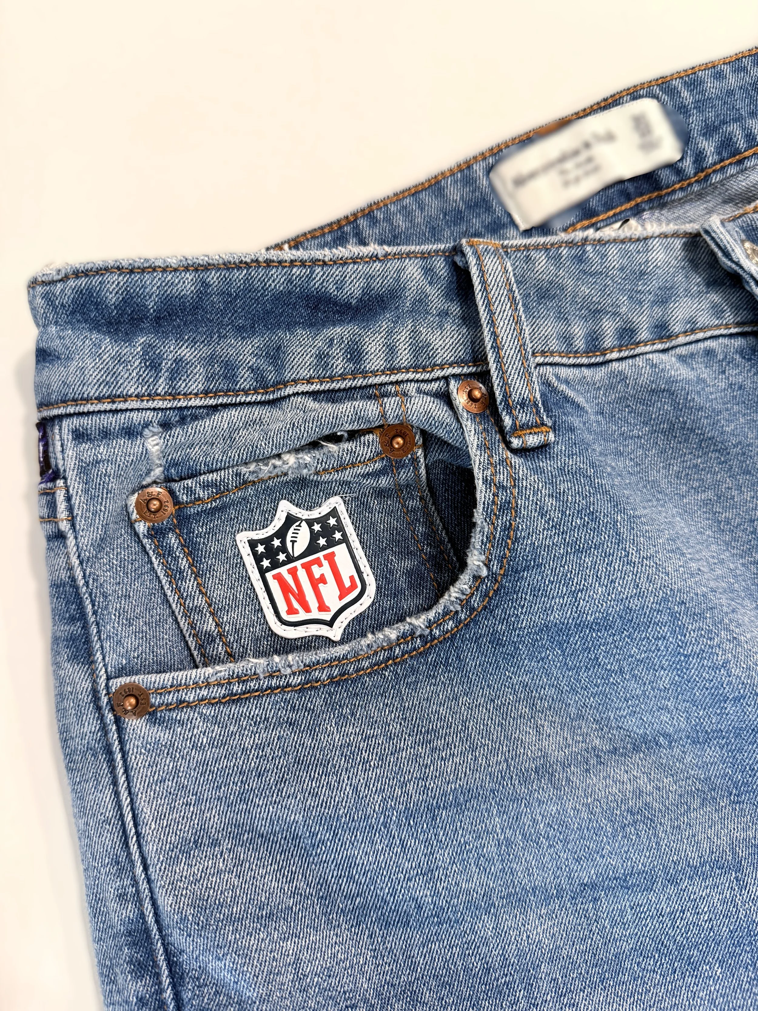 Close-up of a pair of blue denim jeans with an NFL patch on the front pocket and copper rivets.
