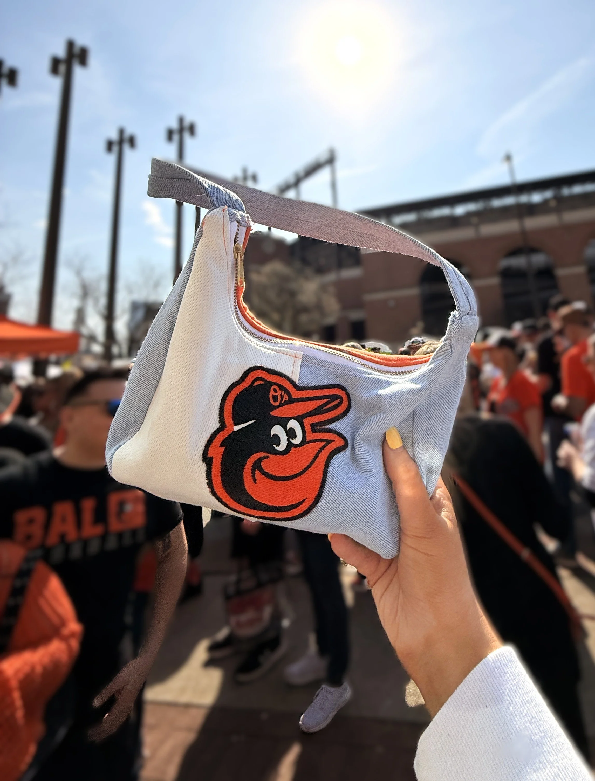 Person holding up a small fabric bag with the Baltimore Orioles logo at a crowded outdoor event on a sunny day.