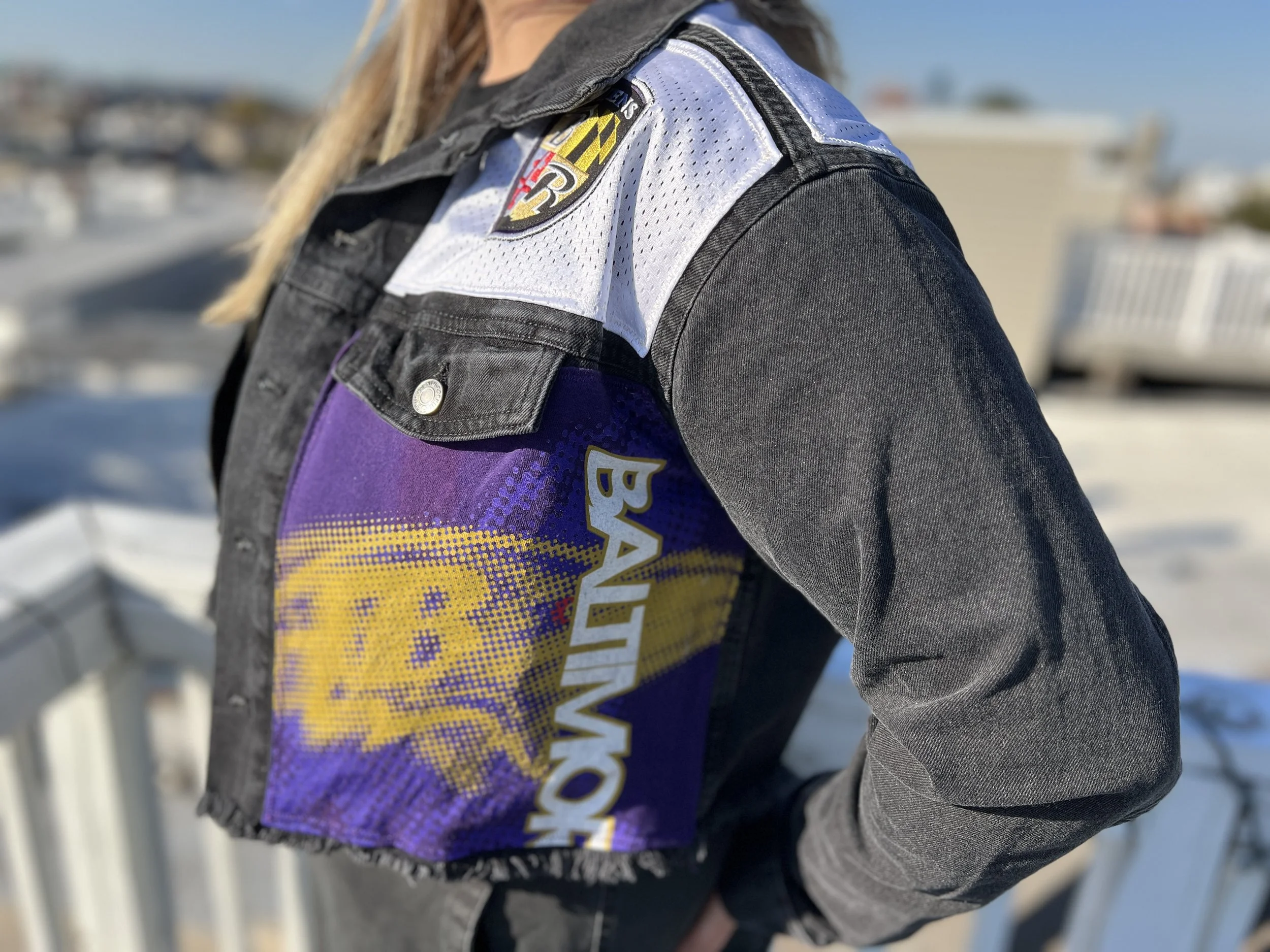 Close-up of a person wearing a denim jacket with patches, including a Baltimore Ravens logo, with a blurred outdoor background.