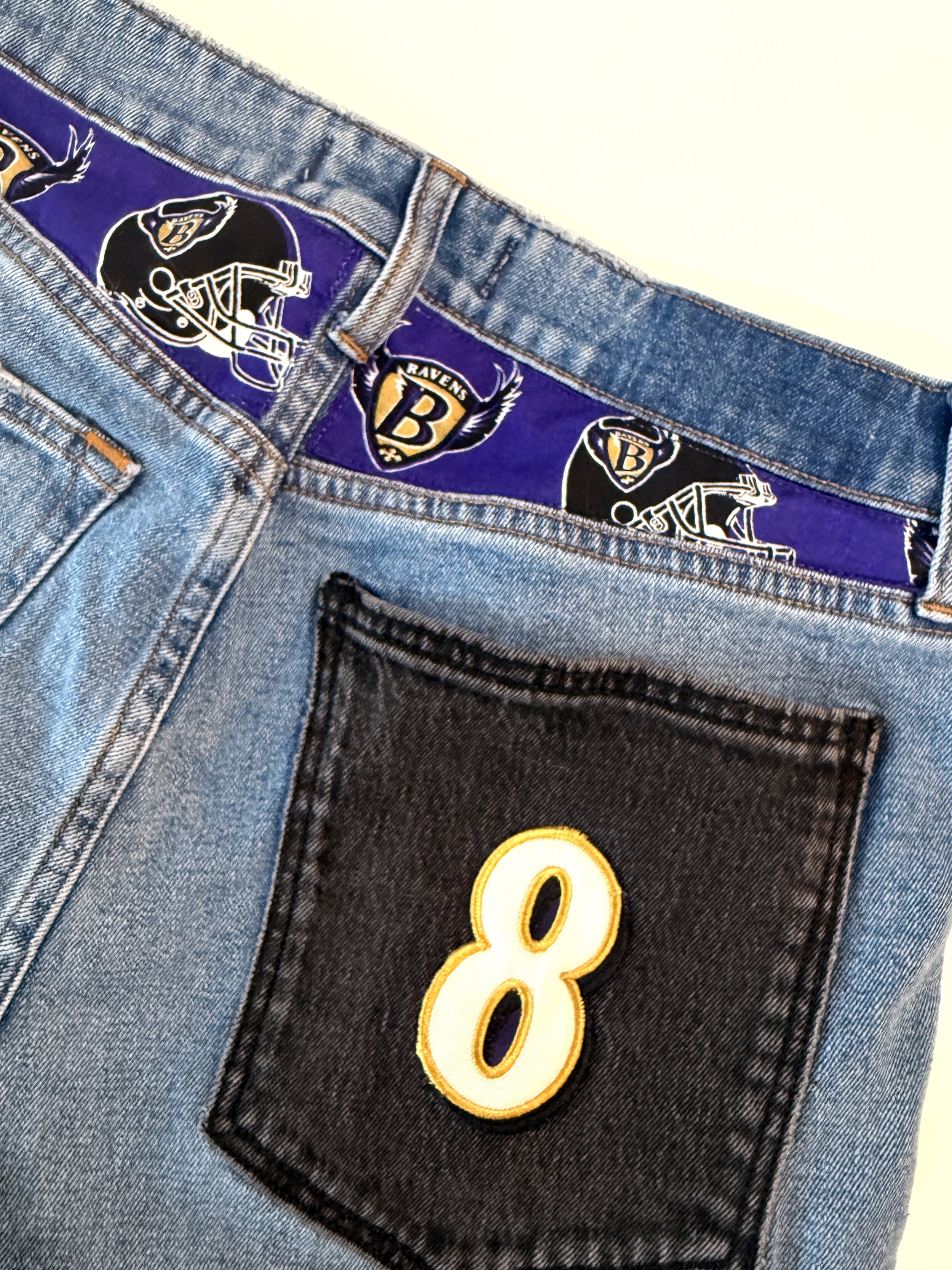 Close-up of denim jeans with a purple fabric tag underneath the waistband, displaying a Baltimore Ravens logo and a large, embroidered number 8 in yellow and black.