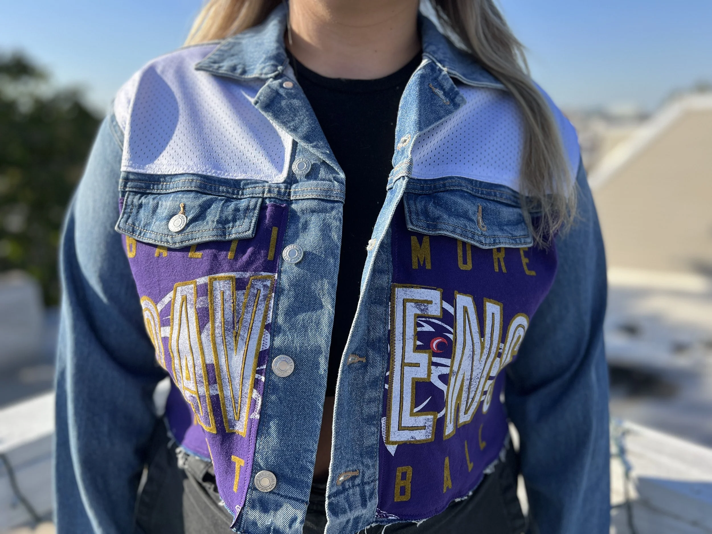 Close-up of a person wearing a denim jacket with a purple sports jersey underneath. The person is outdoors with a blurred background of trees and sky.