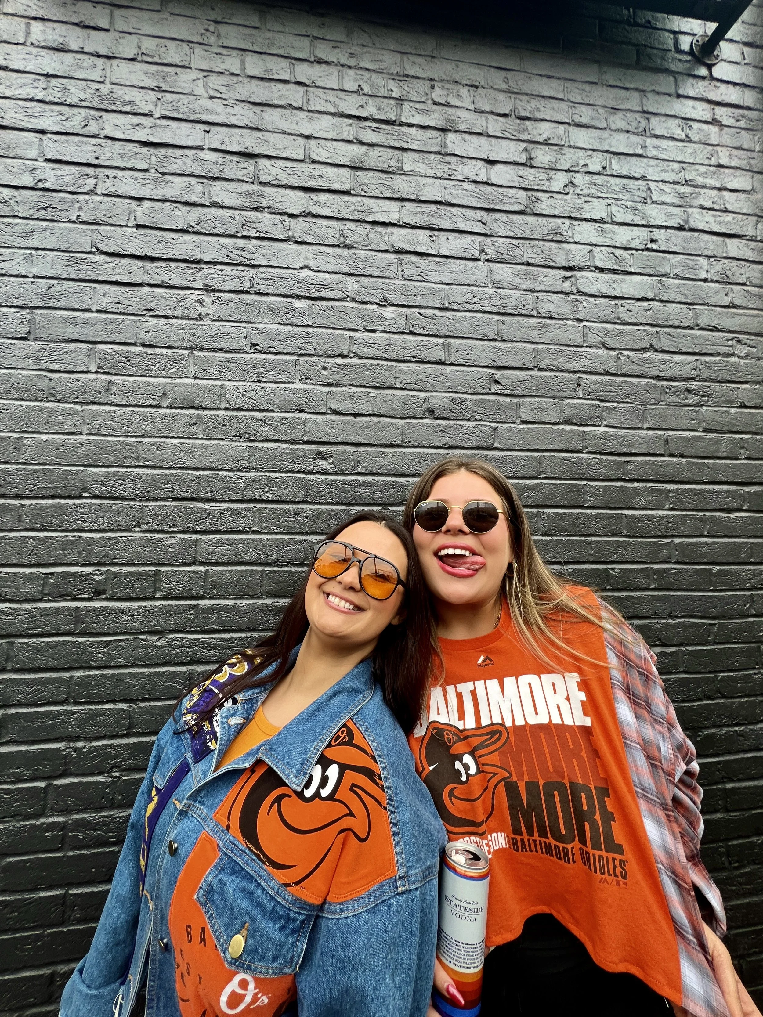 Photo of two women wearing custom Andiamo Designs pieces. One features an orioles denim jacket and the other is wearing an orioles themed t-shirt flannel.