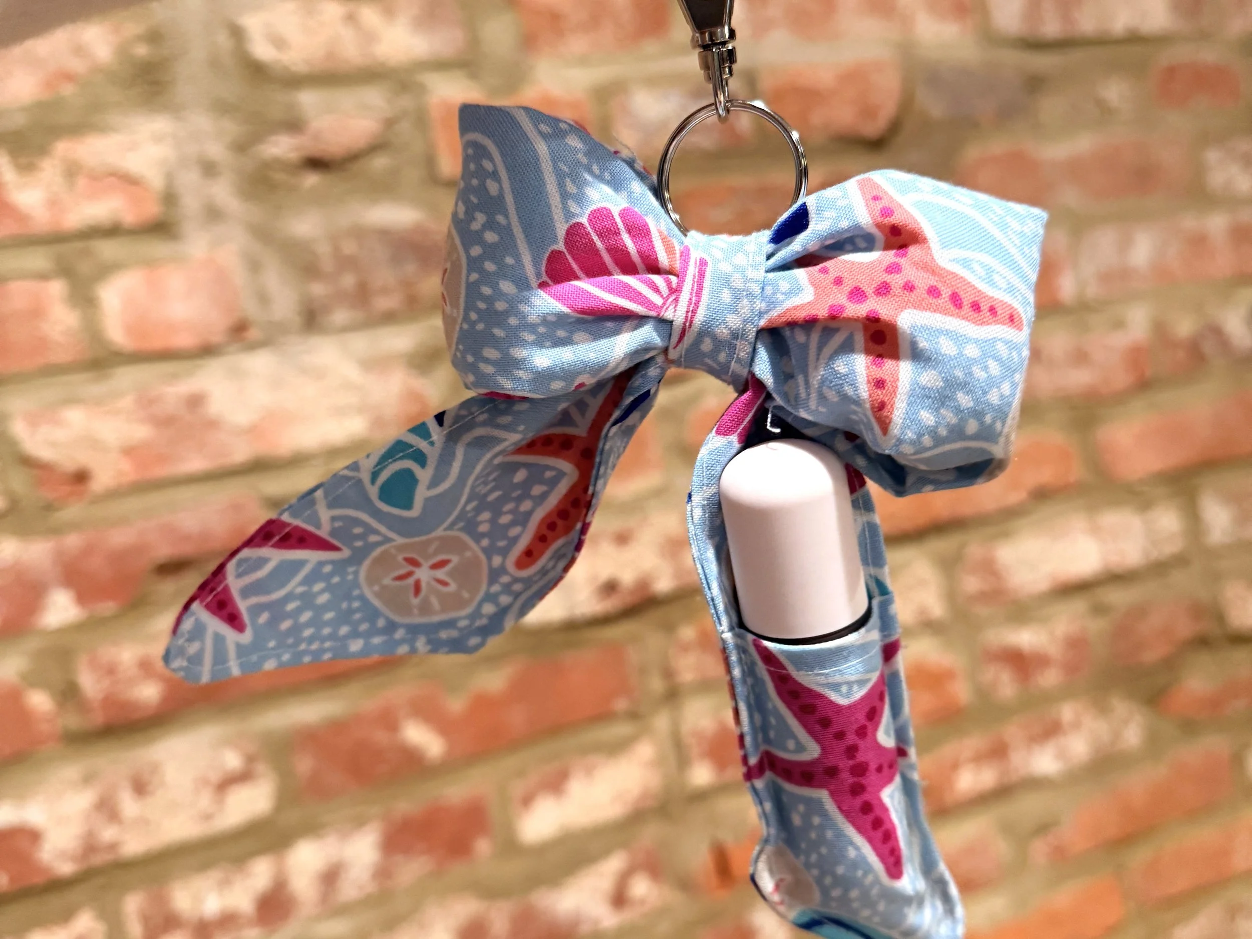 Close-up of a hand holding a hook with a fabric bow attached, against a brick wall background.