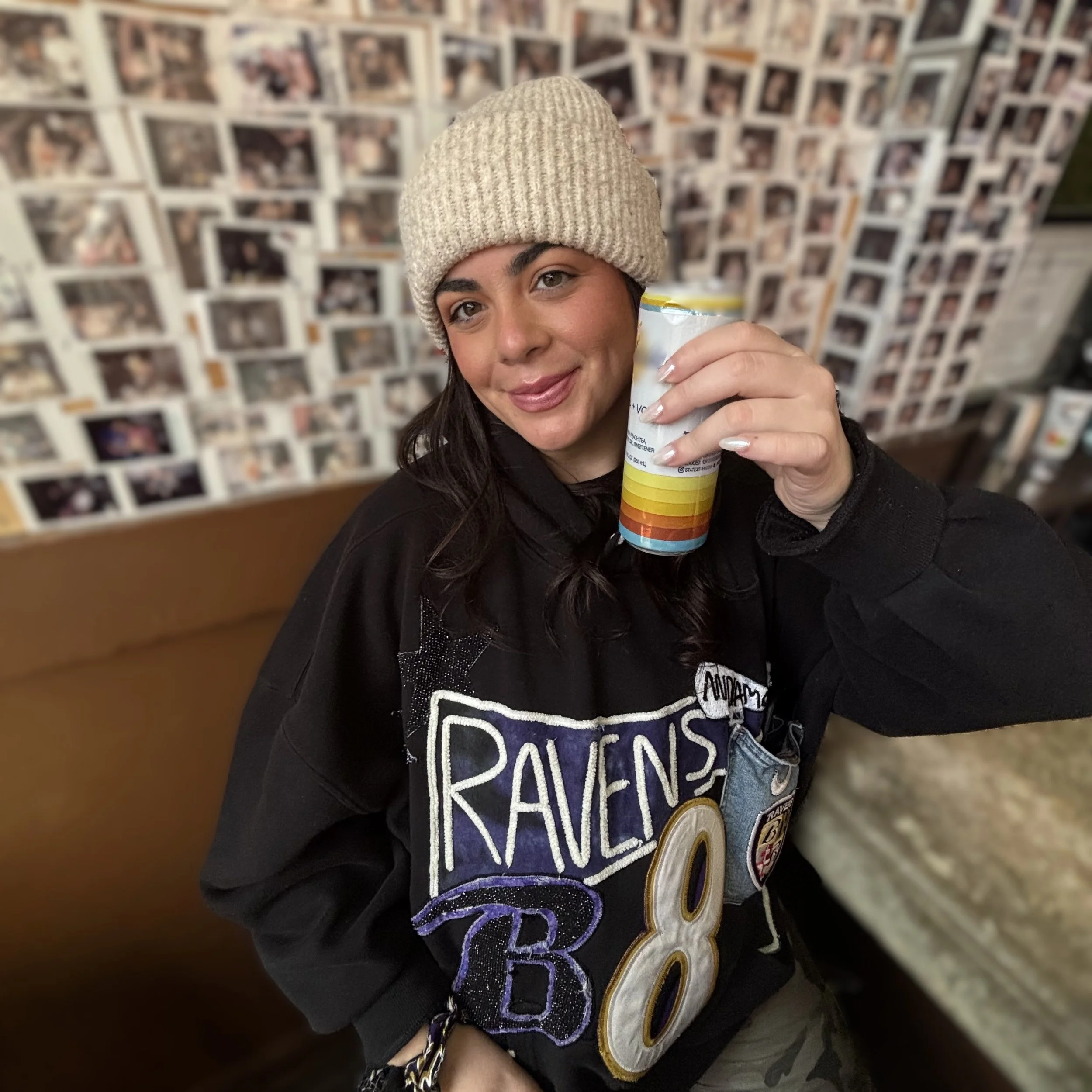 A woman wearing a beige knit beanie, with a custom Ravens hoodie deisgn, smiling at the camera. The background is blurred with a wall of numerous photographs.
