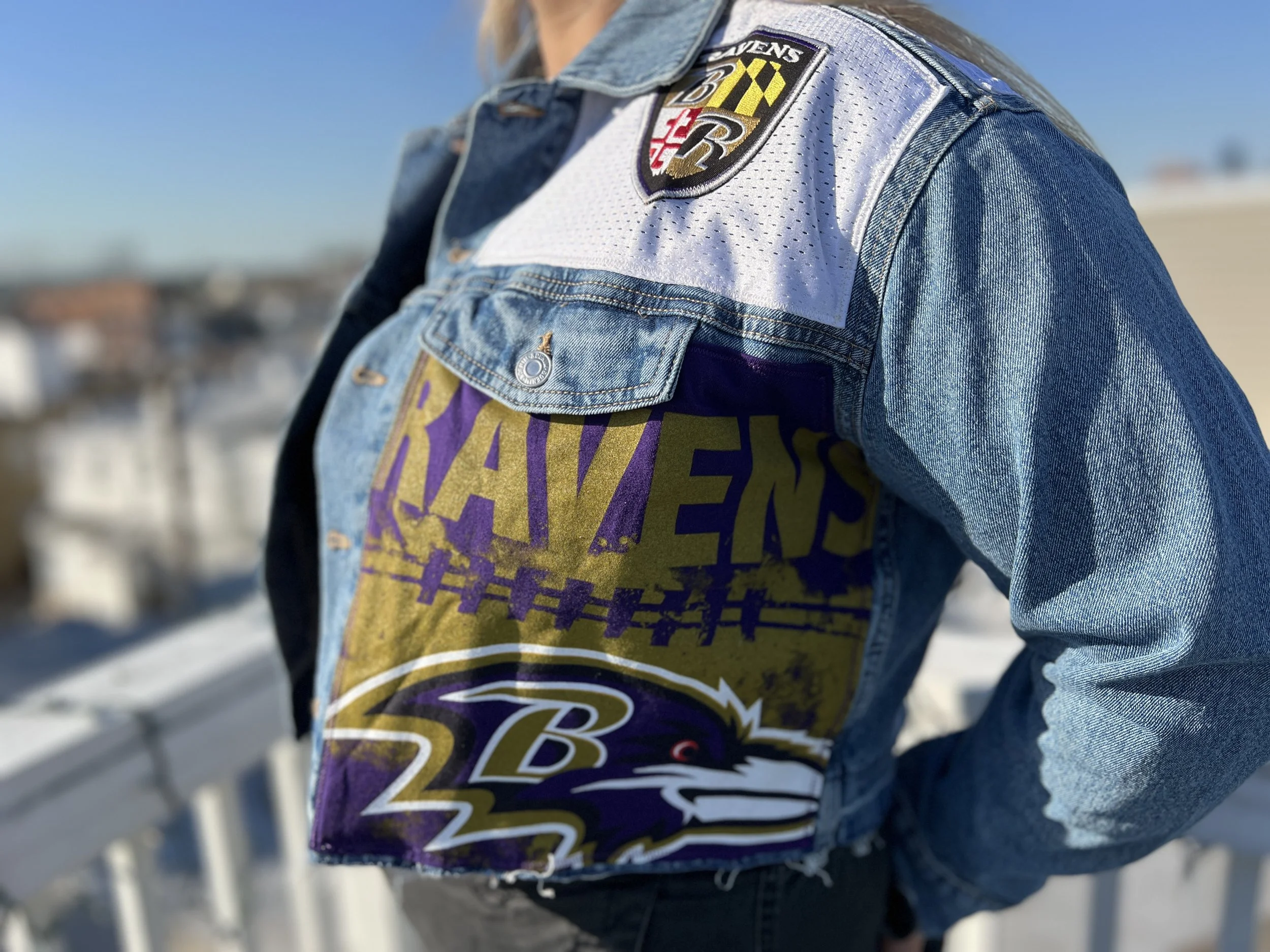 Close-up of a person wearing a Baltimore Ravens NFL team denim jacket with a Ravens patch on the shoulder and t-shirt underneath.