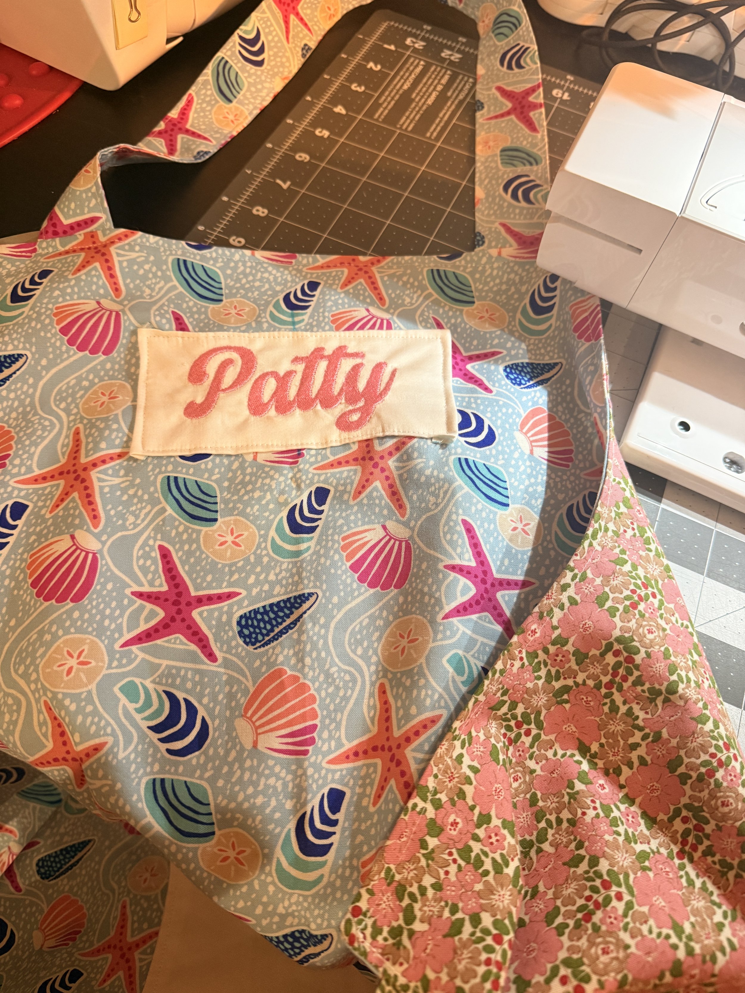 A custom apron folded over to showcase two different patterns, one with a starfish and seashell pattern, and a pink floral cloth, on a table.