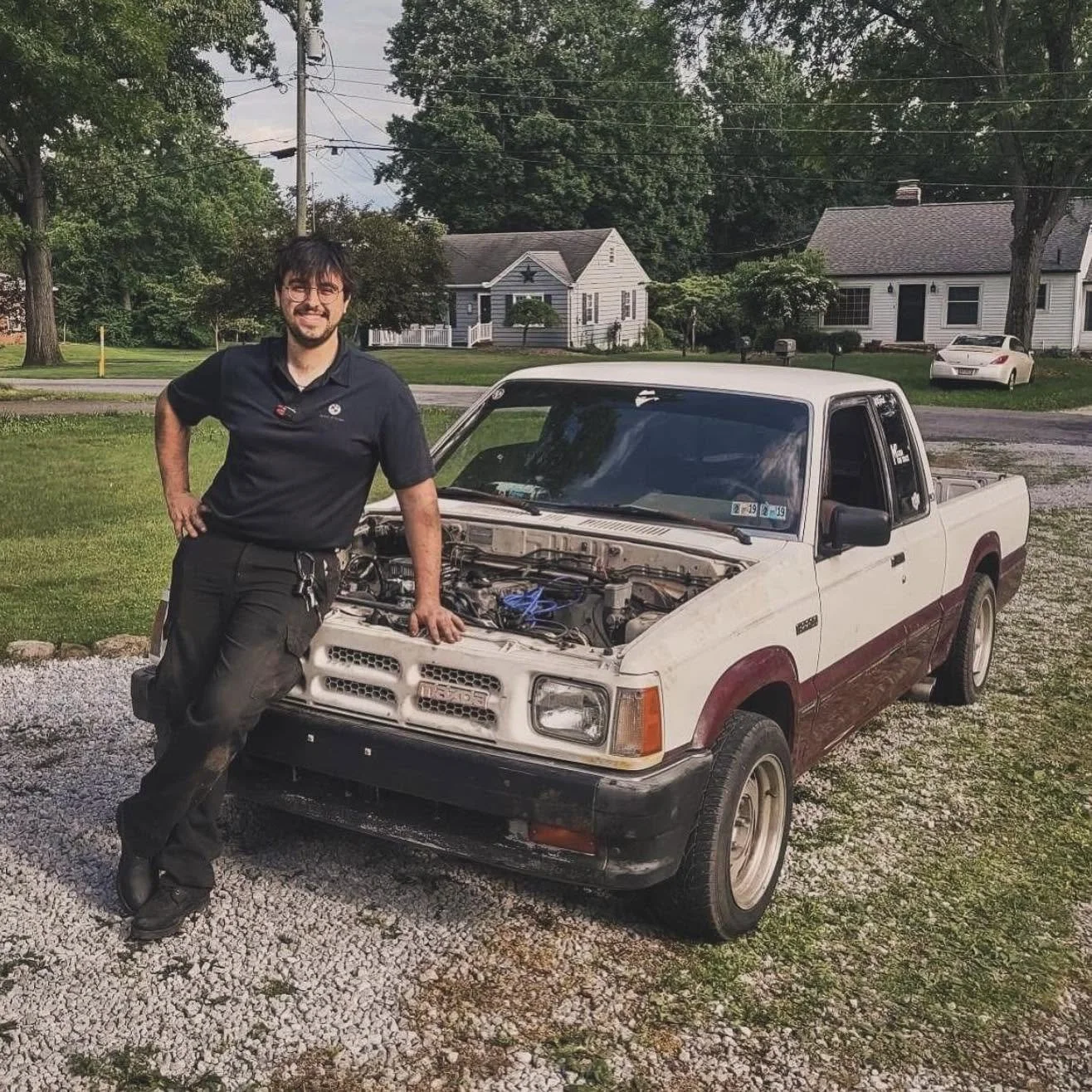 A man with glasses and a beard, wearing black work clothes, is standing next to a white pickup truck with the hood open, in a residential neighborhood with green trees and houses in the background.
