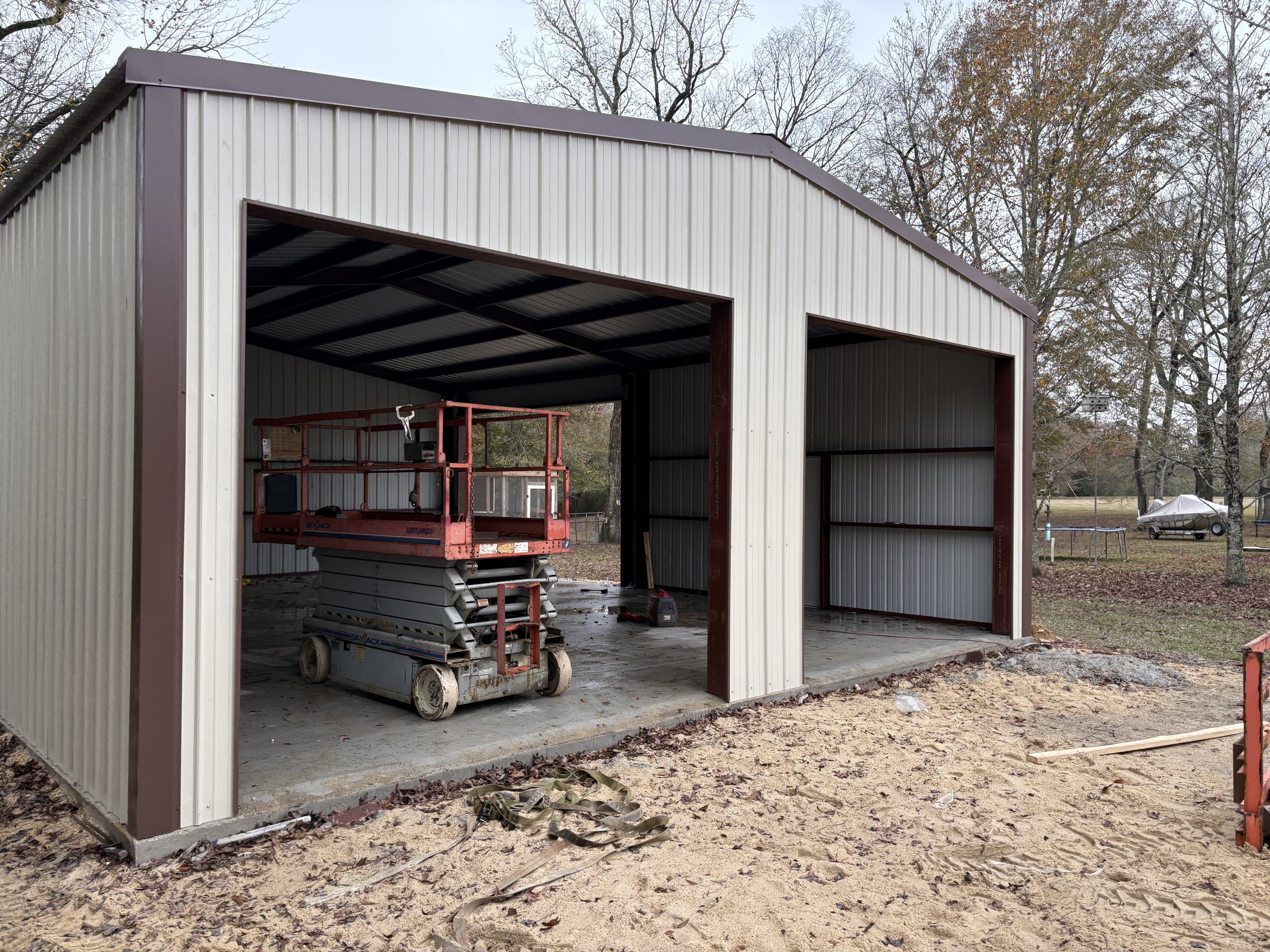Construction or storage building with open front, metal siding, and a scissor lift inside. Trees and a parked car are in the background.