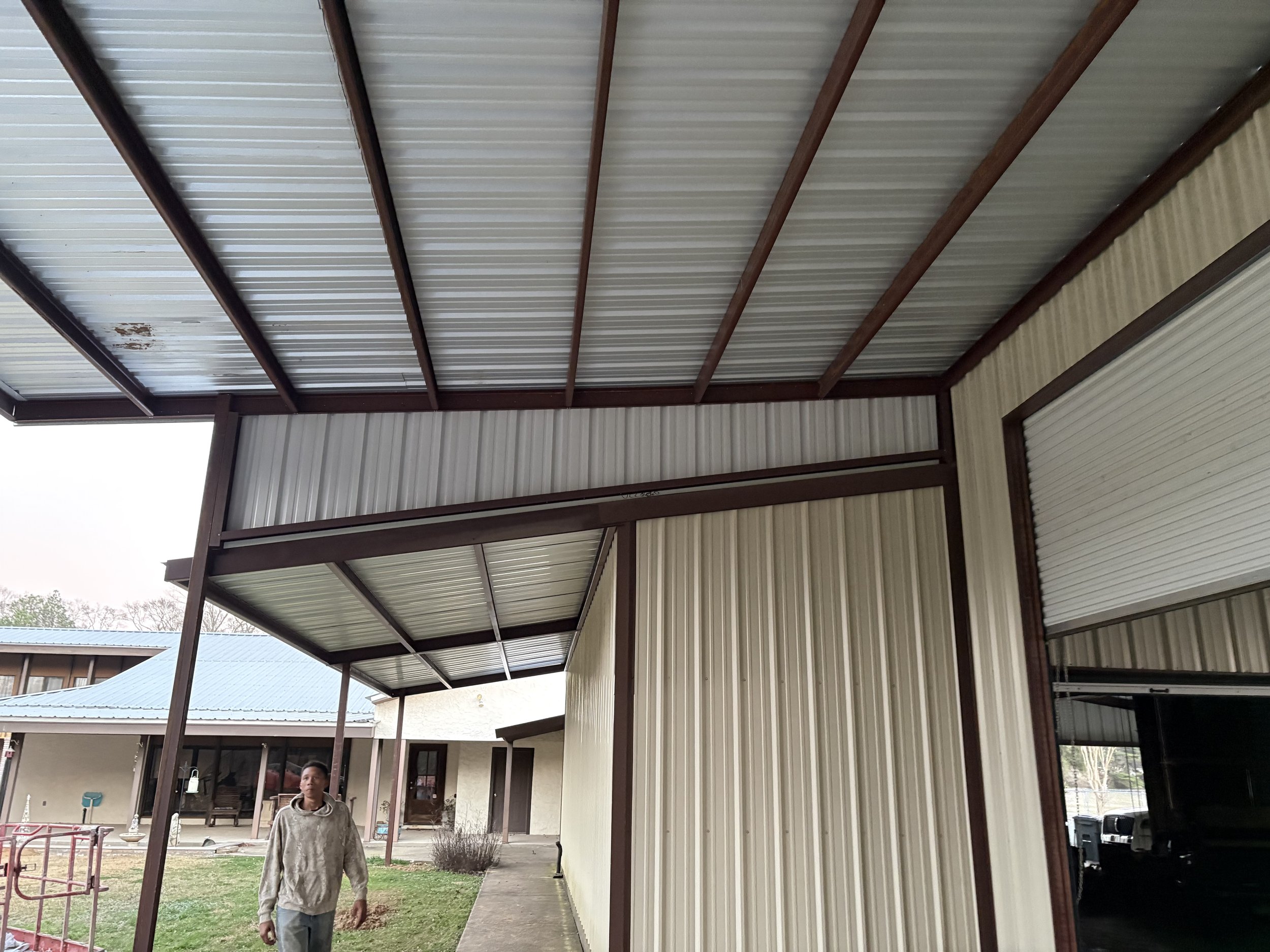 The image shows the exterior of a house with a metal roof and metal siding. There is a covered porch area supported by brown metal beams. A boy wearing gray hoodie and pants is standing on the grass in front of the house, and other houses can be seen in the background.