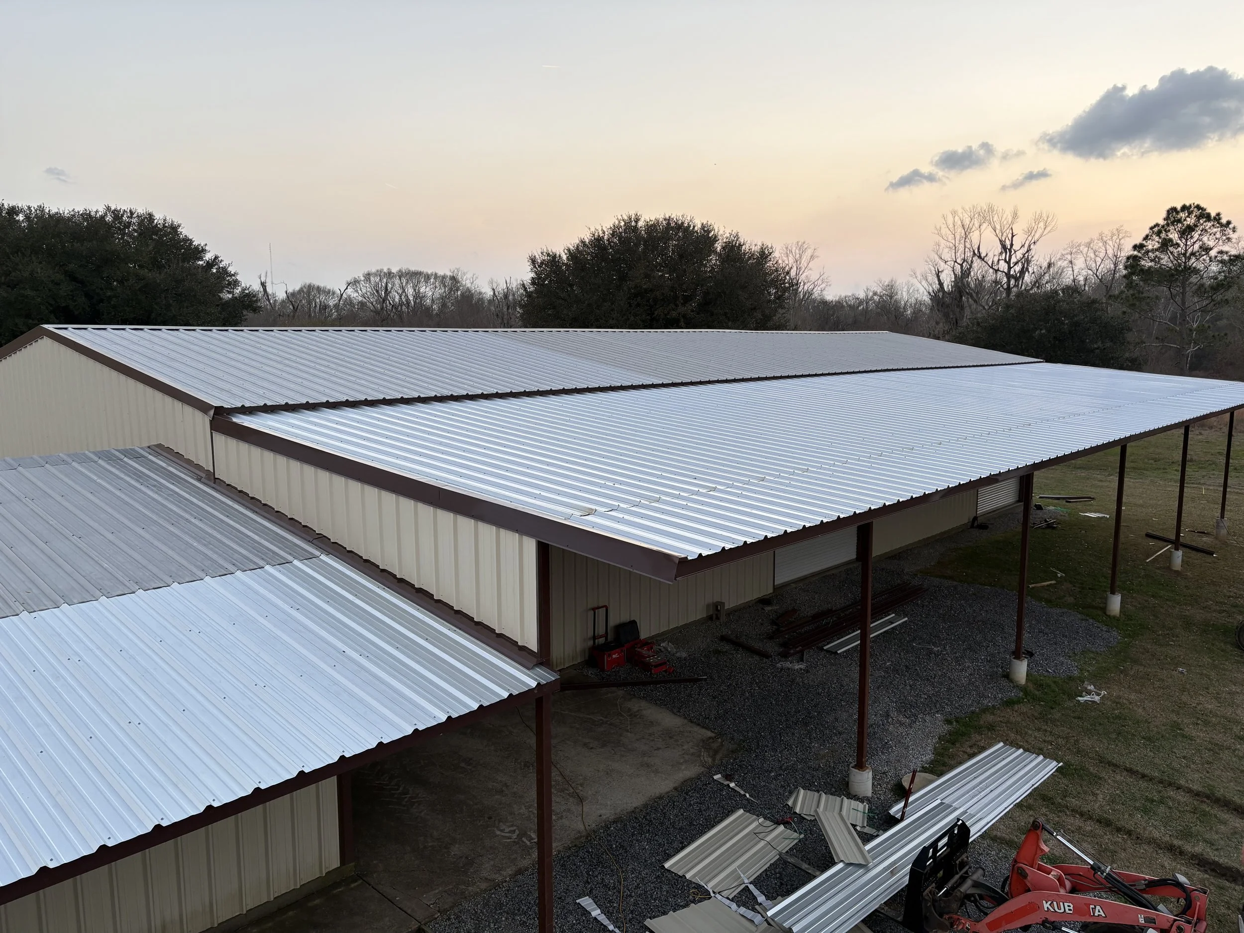 Construction site with metal roofing in progress on large building, surrounded by trees and cloudy sky.