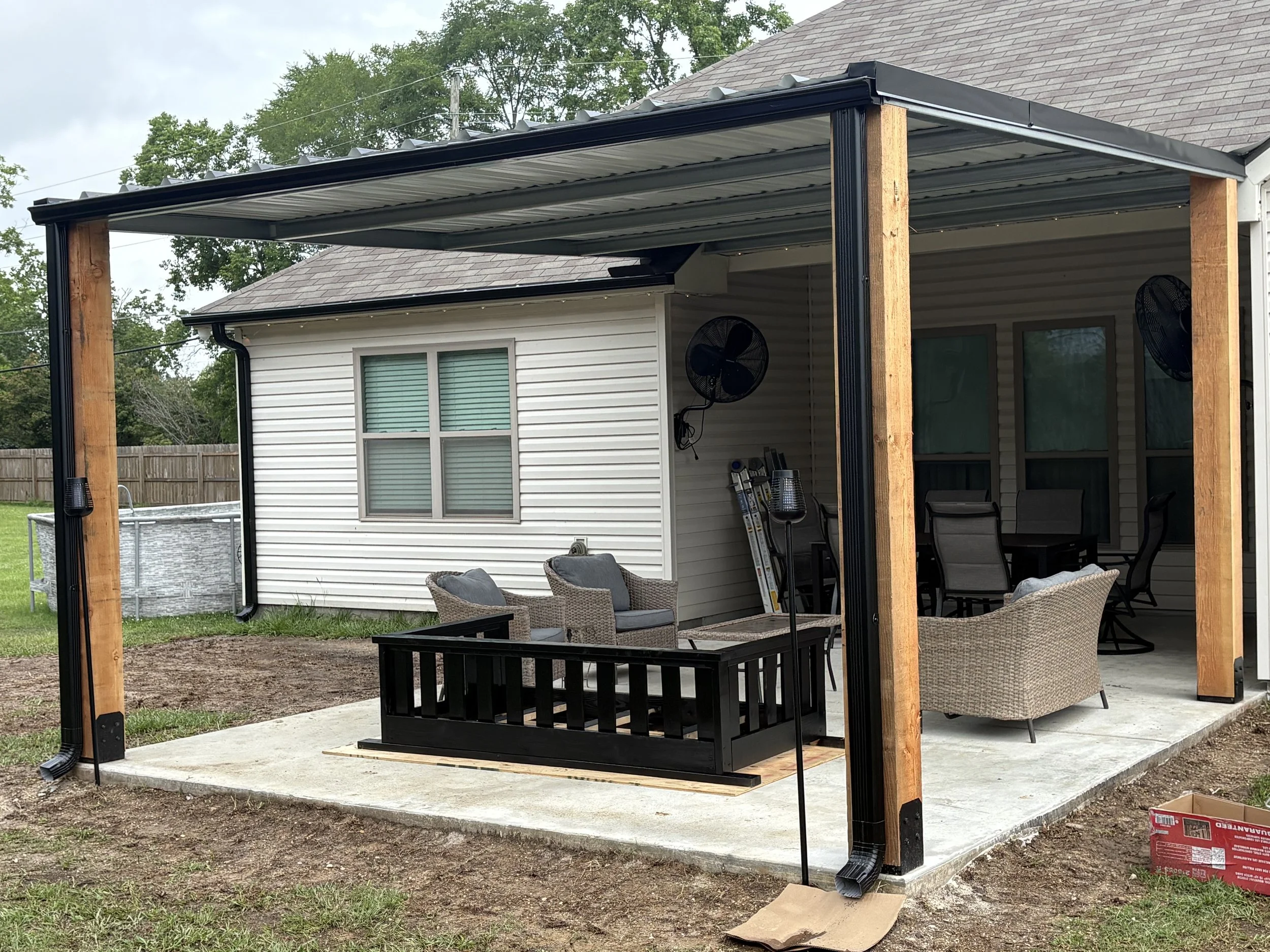 A backyard patio area under construction with a newly built covered patio featuring a metal roof and wooden posts. The patio has outdoor furniture including chairs and a coffee table, and there is a house with white vinyl siding and windows in the background. The ground around the patio is partially bare dirt, and construction materials are visible on the ground.