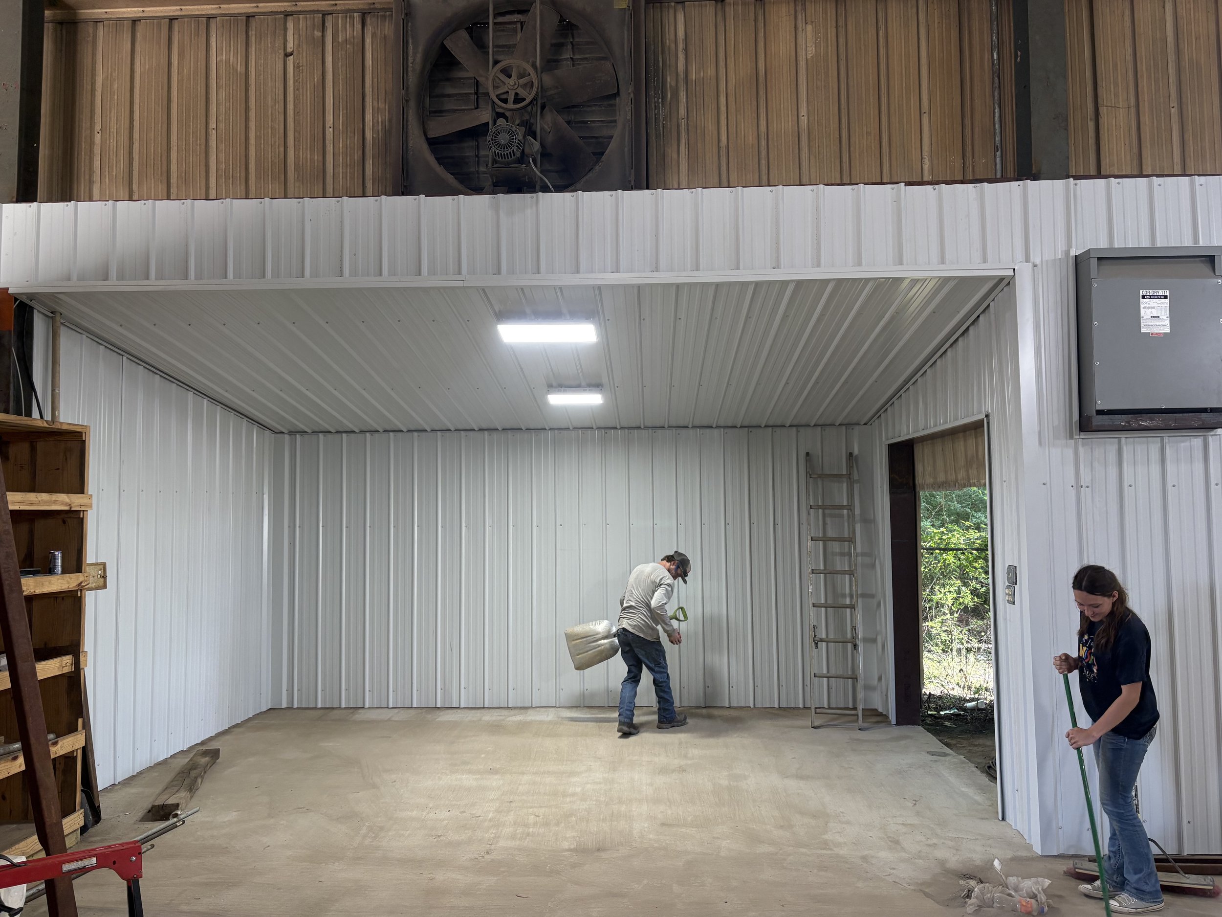 Two people working inside an unfinished building with white metal walls and a wooden ceiling. One person is holding a bag, and the other is cleaning the floor, with sunlight coming in from an open door.