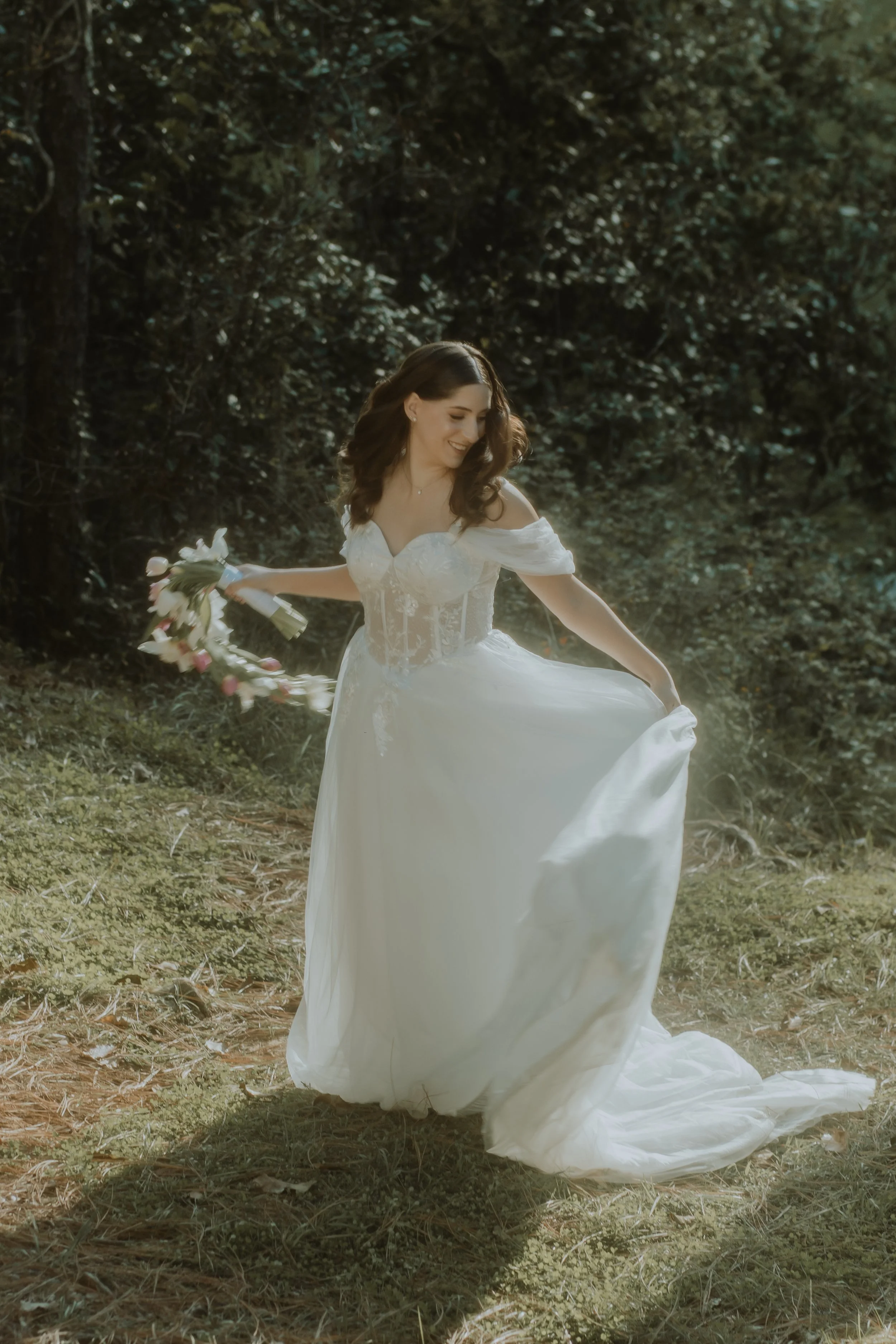 Novia caminando entre árboles con vestido blanco y ramo de flores