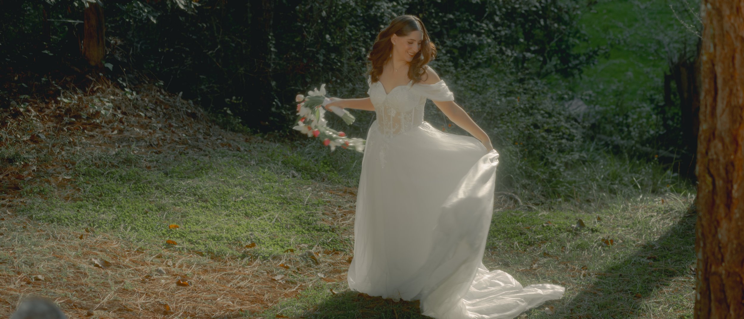 Novia caminando entre árboles con vestido blanco y ramo de flores