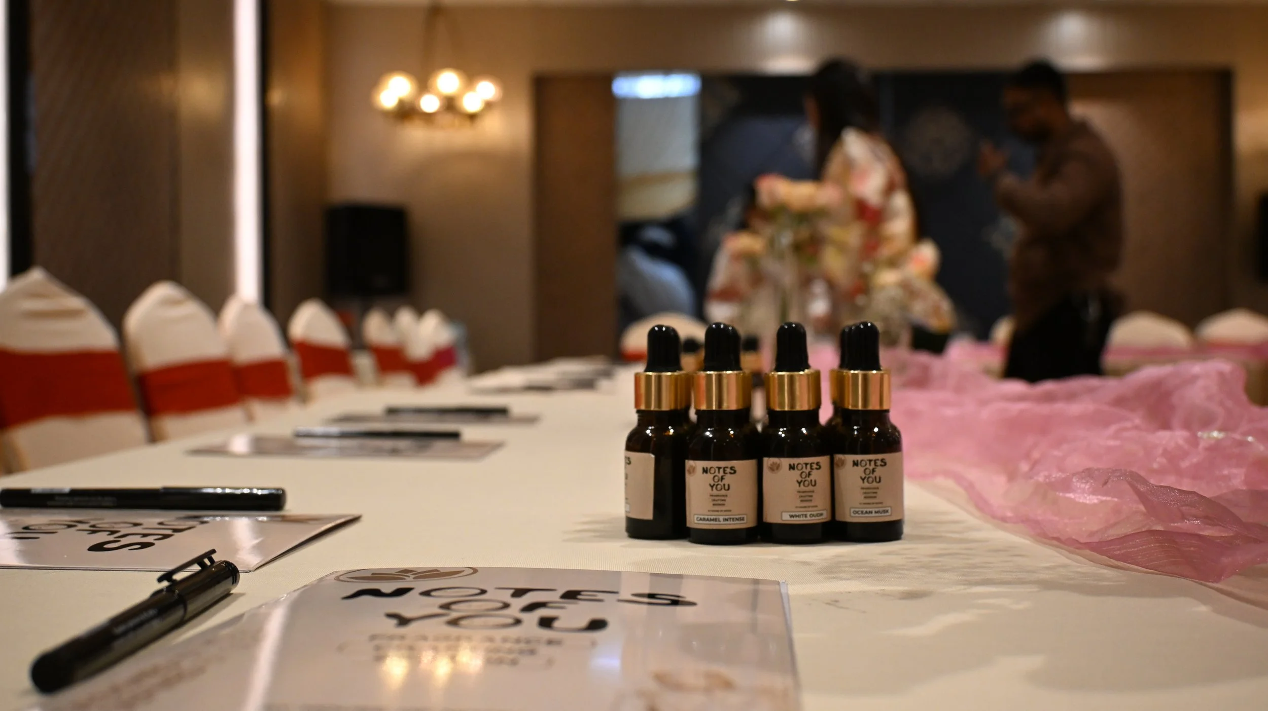 Five small bottles with droppers labeled with different scents or flavors, arranged on a table with a pink cloth, with chairs and people in the background