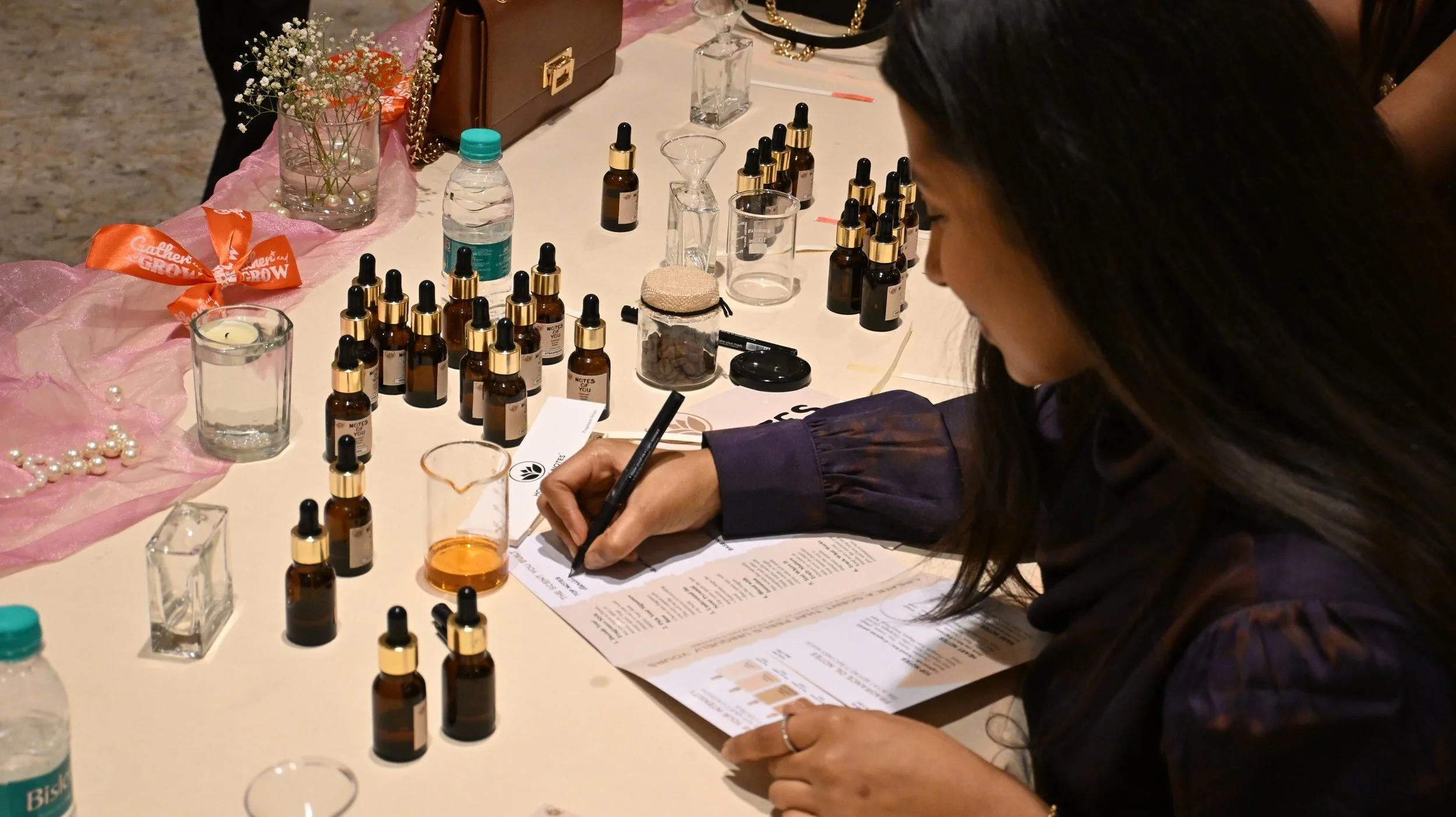 A woman writing on a sheet of paper at a table, surrounded by small brown bottles with dropper caps, cups, a water bottle, and decorative items like flowers and pearl necklaces.
