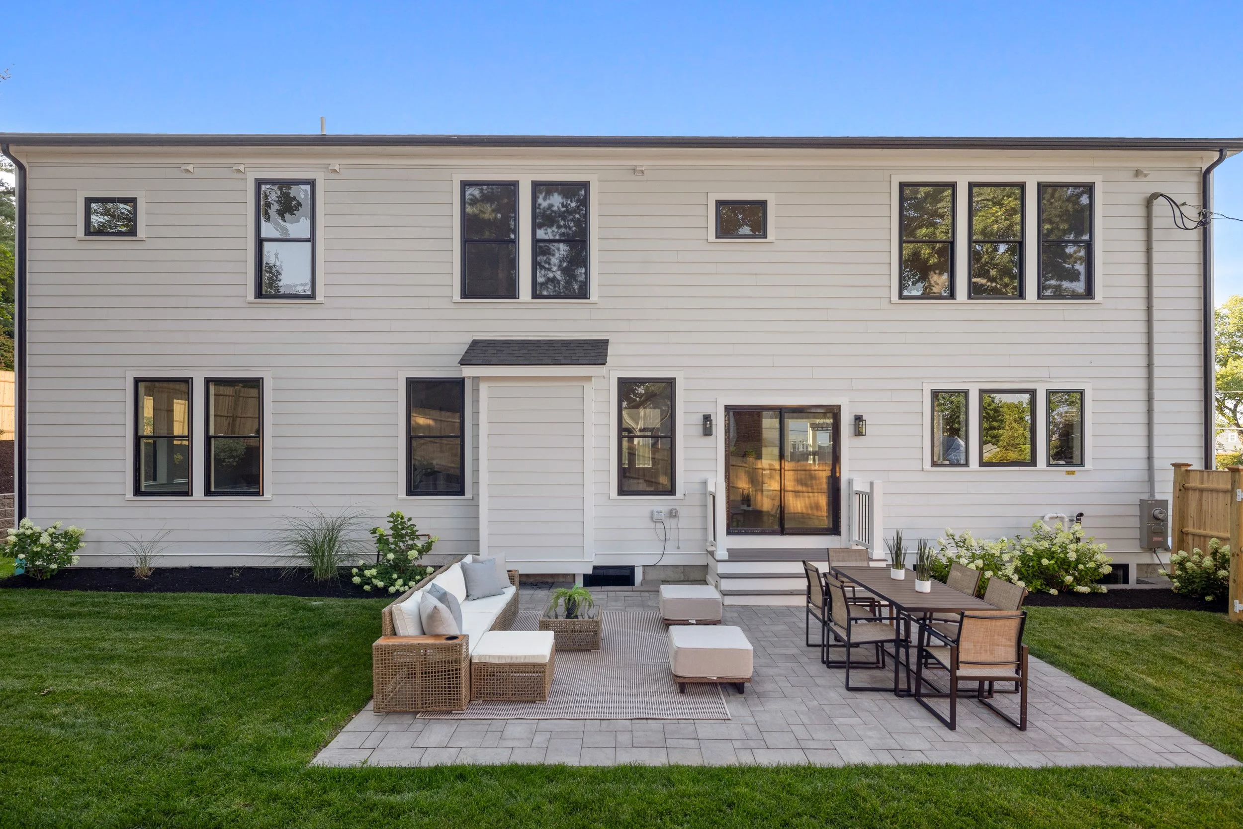 Backyard patio with outdoor furniture including a wicker sofa, white cushions, and a dining table with chairs, all on a stone-paved area. There's a well-maintained lawn with small plants in front of a white two-story house with black-framed windows, 