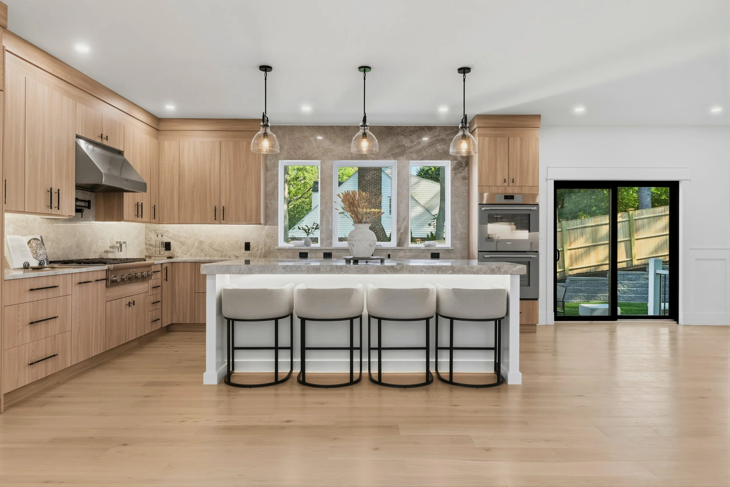Modern kitchen with light wood cabinets, marble countertop, three pendant lights over a central island, large window with views of greenery, sliding glass door, and a cream-colored barstool at the island.