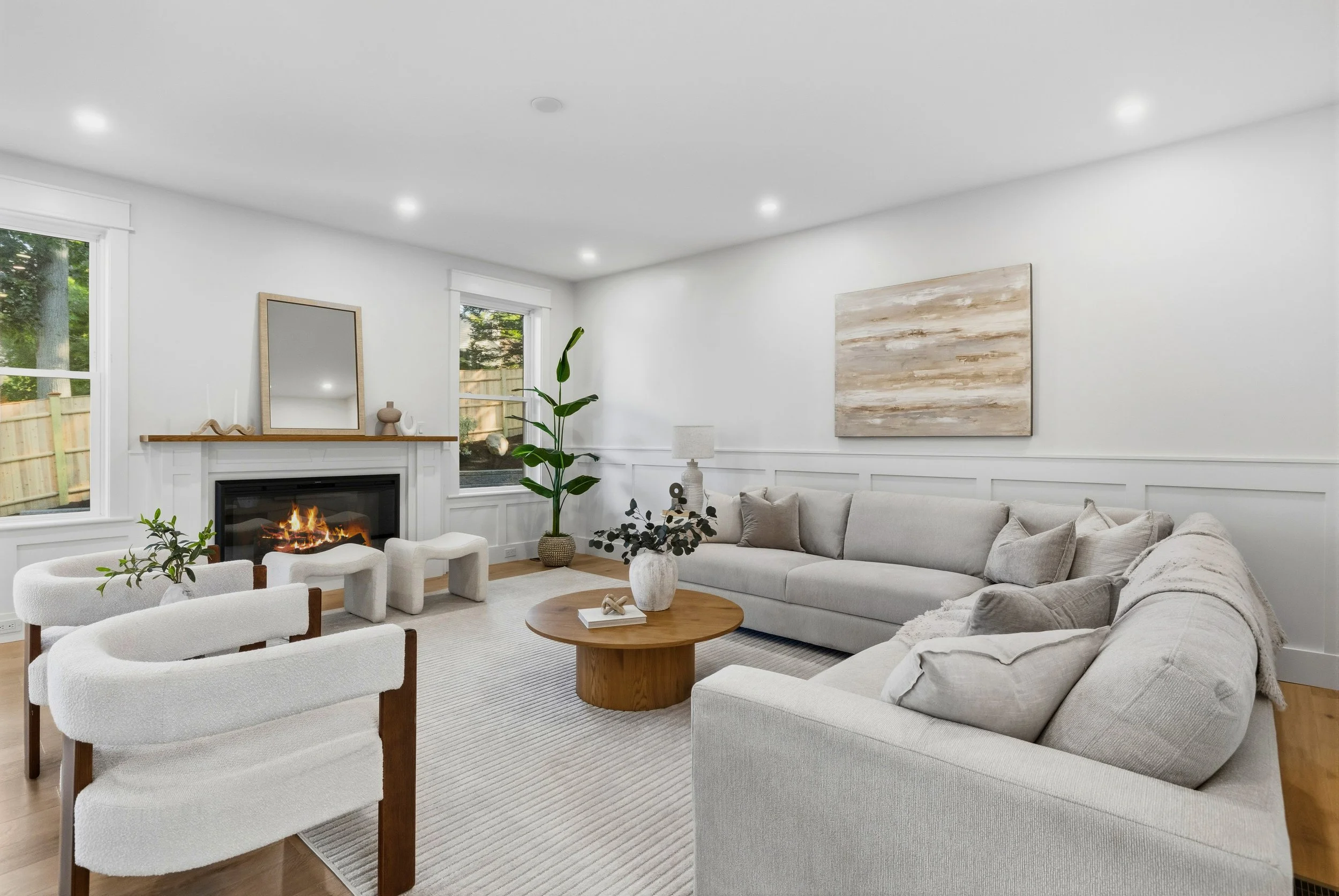 Bright living room with white walls, large window, gray sectional sofa, white armchairs, round wooden coffee table, fireplace with mirror above, potted plant, and abstract artwork on the wall.