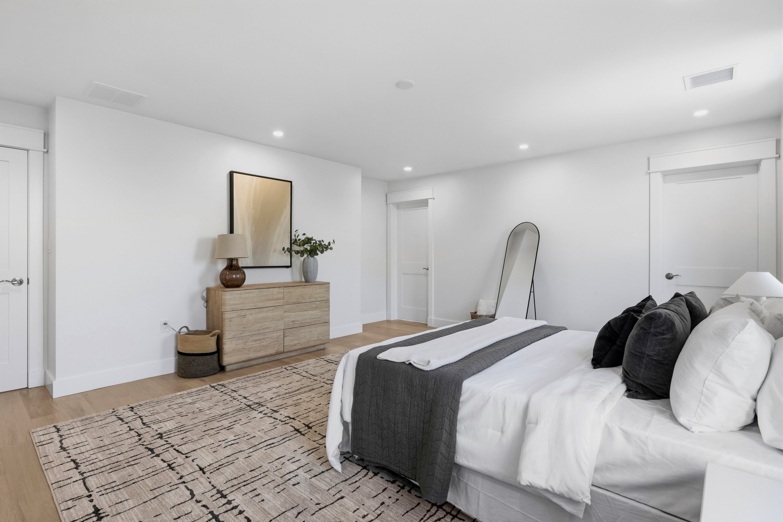 A modern bedroom with white walls and ceiling, a bed with white and black bedding, a wooden dresser with decorative items, and a black-framed mirror. There are three doors, a rug on the light wooden floor, and modern ceiling lights.
