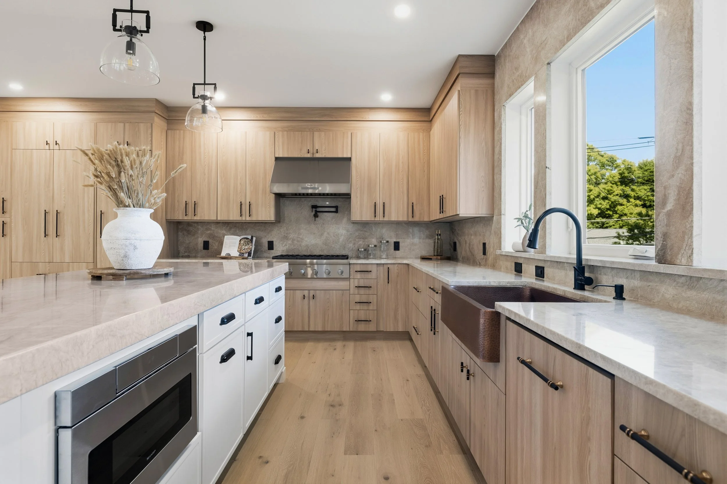 Modern kitchen with light wood cabinets, white lower cabinets, black hardware, stainless steel appliances, marble countertops, and a large window showing green trees outside.
