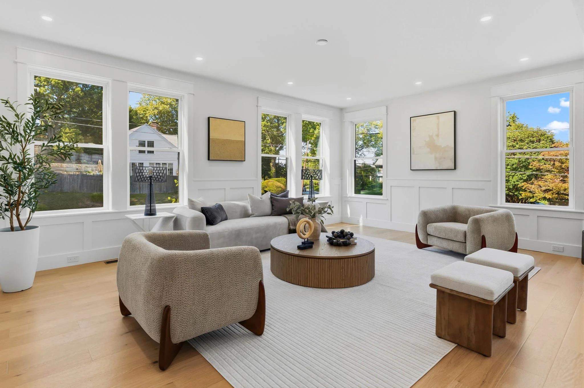 Bright living room with large windows, white walls, beige armchairs, a white sofa, and a round wooden coffee table. Artwork hangs on the walls, and trees can be seen outside.