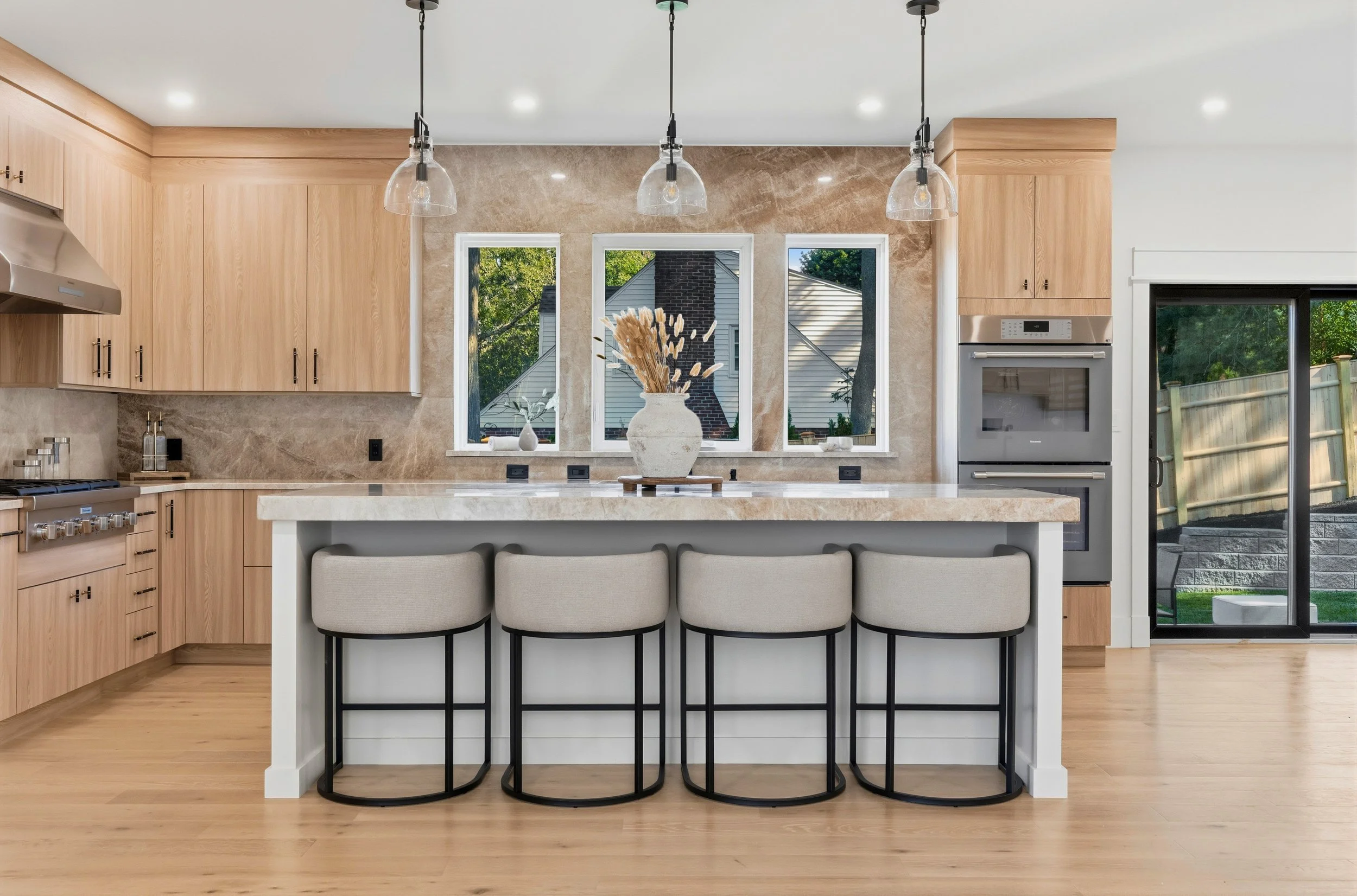 Modern kitchen with light wood cabinets, marble backsplash, and a white kitchen island with four beige upholstered barstools. Pendant lights hang over the island, and a window displays a backyard scene with trees and a house.