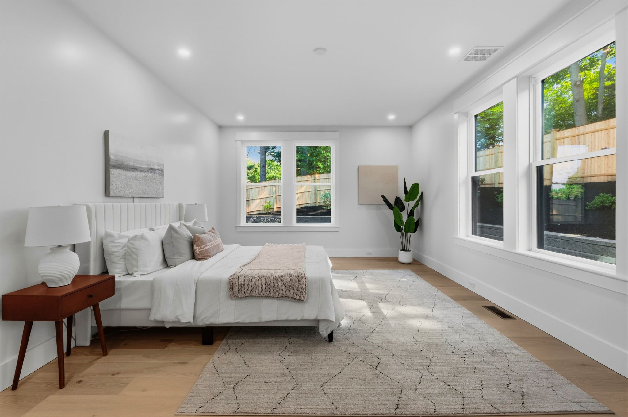 A bright, modern living room with white walls, large windows, a white sofa with pillows, a small wooden side table with a lamp, a potted plant, and a patterned rug on light wooden floor.