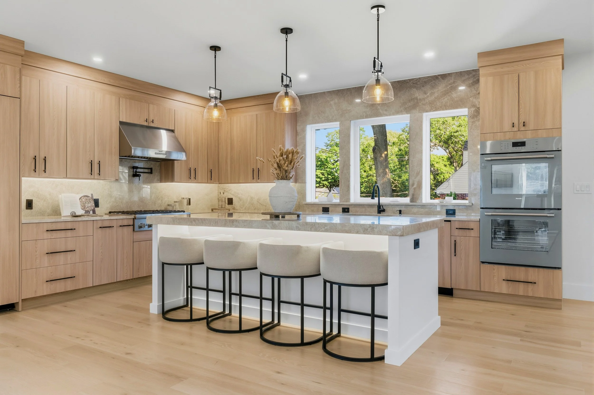 Modern kitchen with light wood cabinets, a white island with a beige marble top, three black bar stools with white cushions, a large vase with dried plants, three pendant lights, and three large windows showing trees outside.