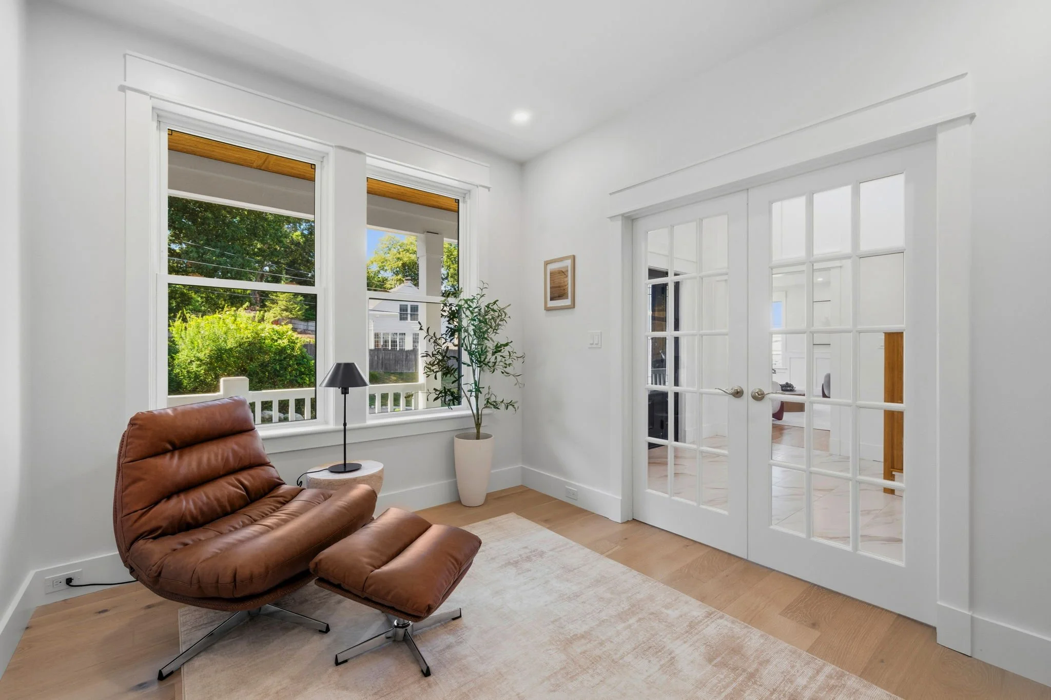 Living room interior with large windows showing greenery outside, a modern brown leather chaise lounge, a black table lamp on a white side table, a potted plant, and white walls with a wooden-framed artwork. French doors leading to another room.
