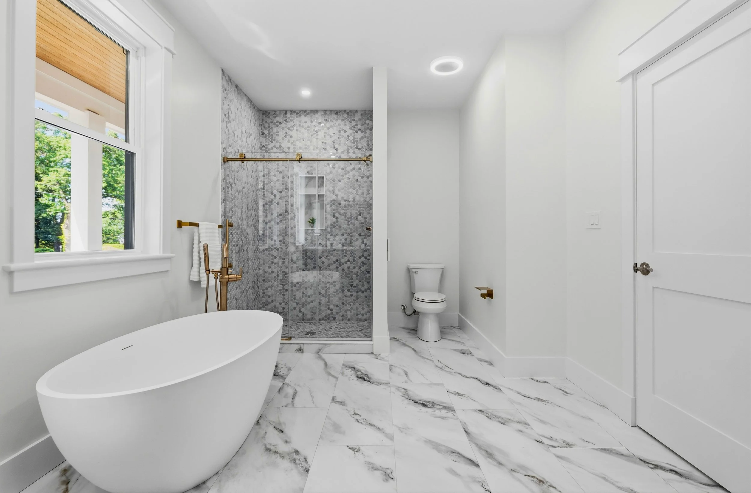 Modern bathroom with white walls, marble flooring, a freestanding bathtub, a window with greenery outside, a shower with grey mosaic tiles and brass fixtures, a white toilet, and a minimalist design.