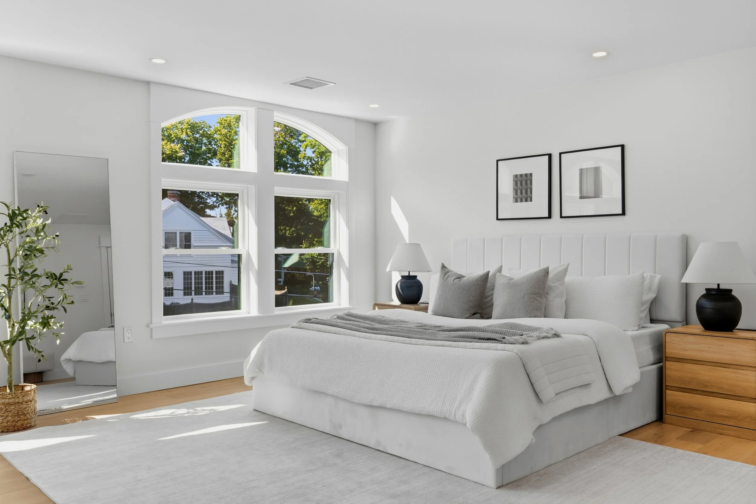 A bright, modern bedroom with large arched windows showing green trees outside. The room features a white bed with gray and white pillows, two black lamps on wooden nightstands, framed abstract artwork on the wall, and a full-length mirror near a pot