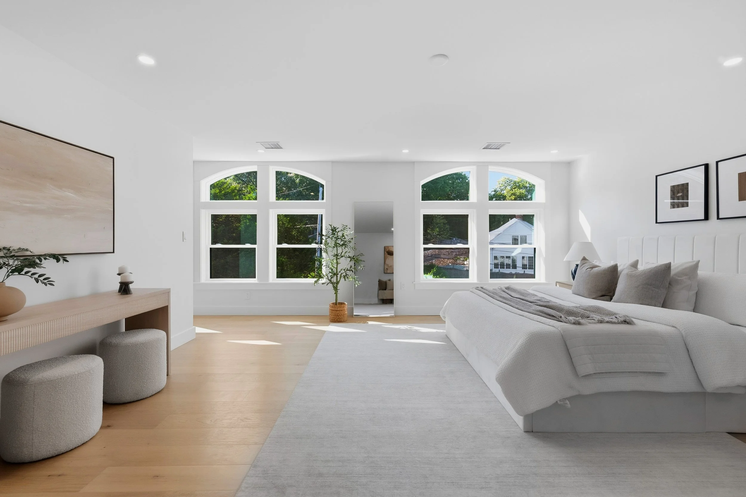 Bright, minimalist bedroom with white walls, large arched windows, a white bed with gray pillows, a gray rug, a potted plant, and framed artwork on the wall.