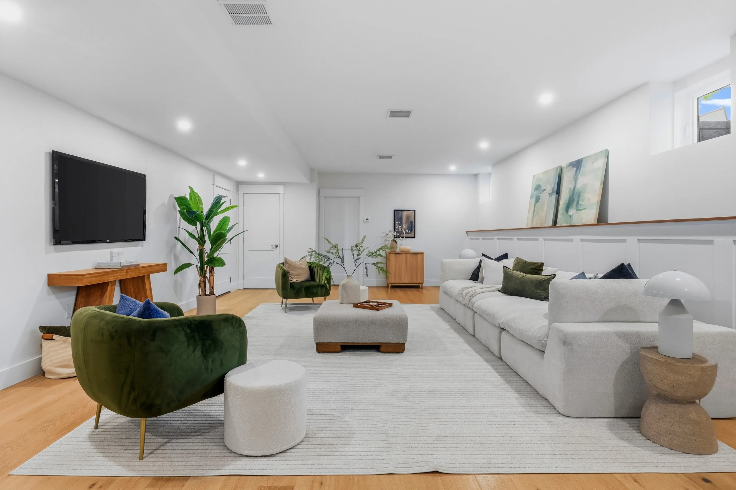 Modern living room with white walls, a large flat-screen TV, green and white armchairs, a white sectional sofa with throw pillows, a wooden side table, abstract paintings, indoor plants, and a light-colored rug.