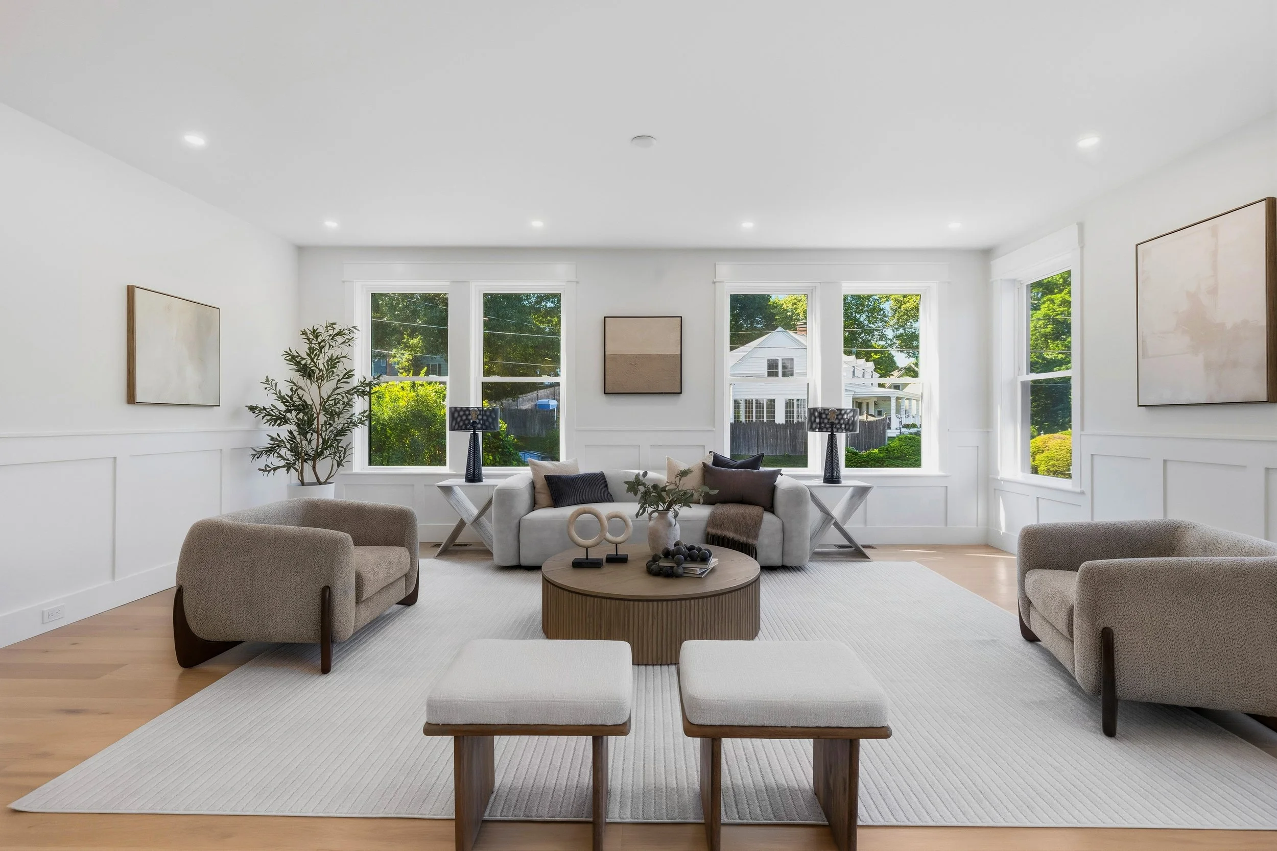 Bright living room with large windows overlooking greenery, a white sofa with pillows, dark lamps, wall art, potted plant, ottomans, and a round coffee table on a white rug.