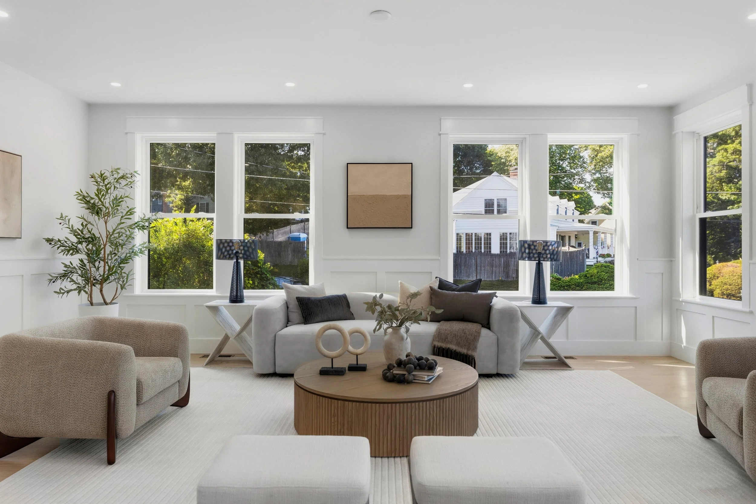 Bright living room with large windows, white walls, white sofa, beige armchair, round wooden coffee table, decorative plants, and artwork on the wall.
