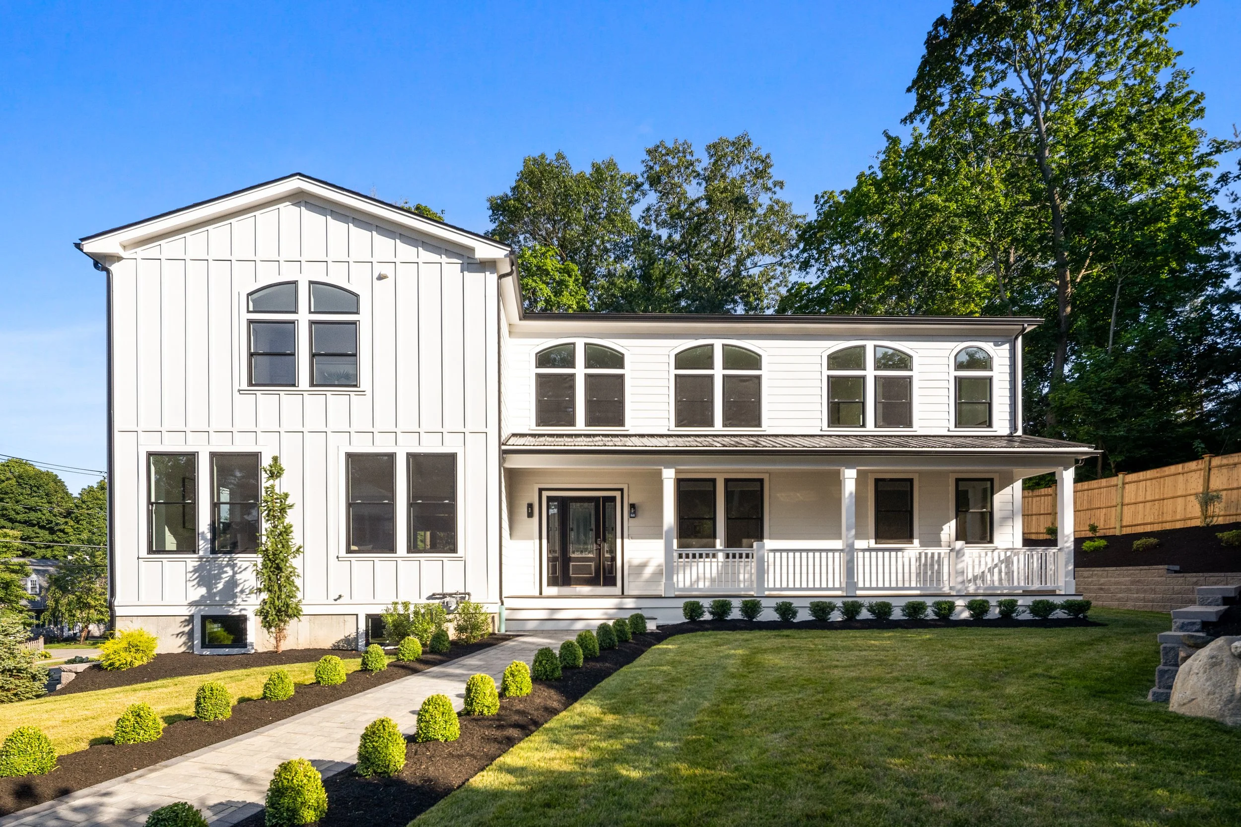 Front view of a white modern house with multiple windows, a porch, and a landscaped yard with a walkway, small bushes, and a green lawn, with trees and a clear blue sky in the background.