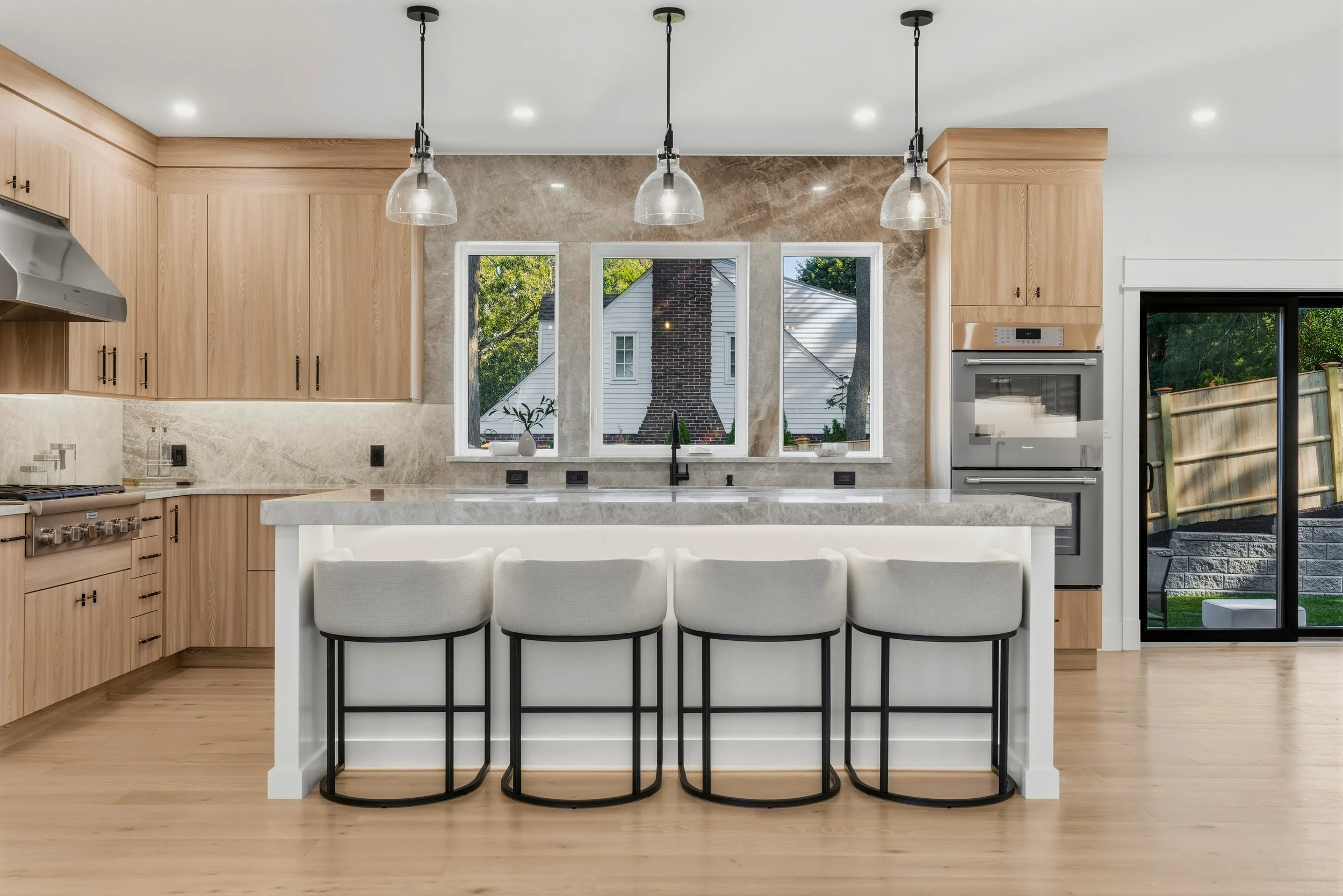 Modern kitchen with light wood cabinets, white island, three black and white bar stools, marble backsplash, three pendant lights, large window with view of backyard, and stainless steel appliances.