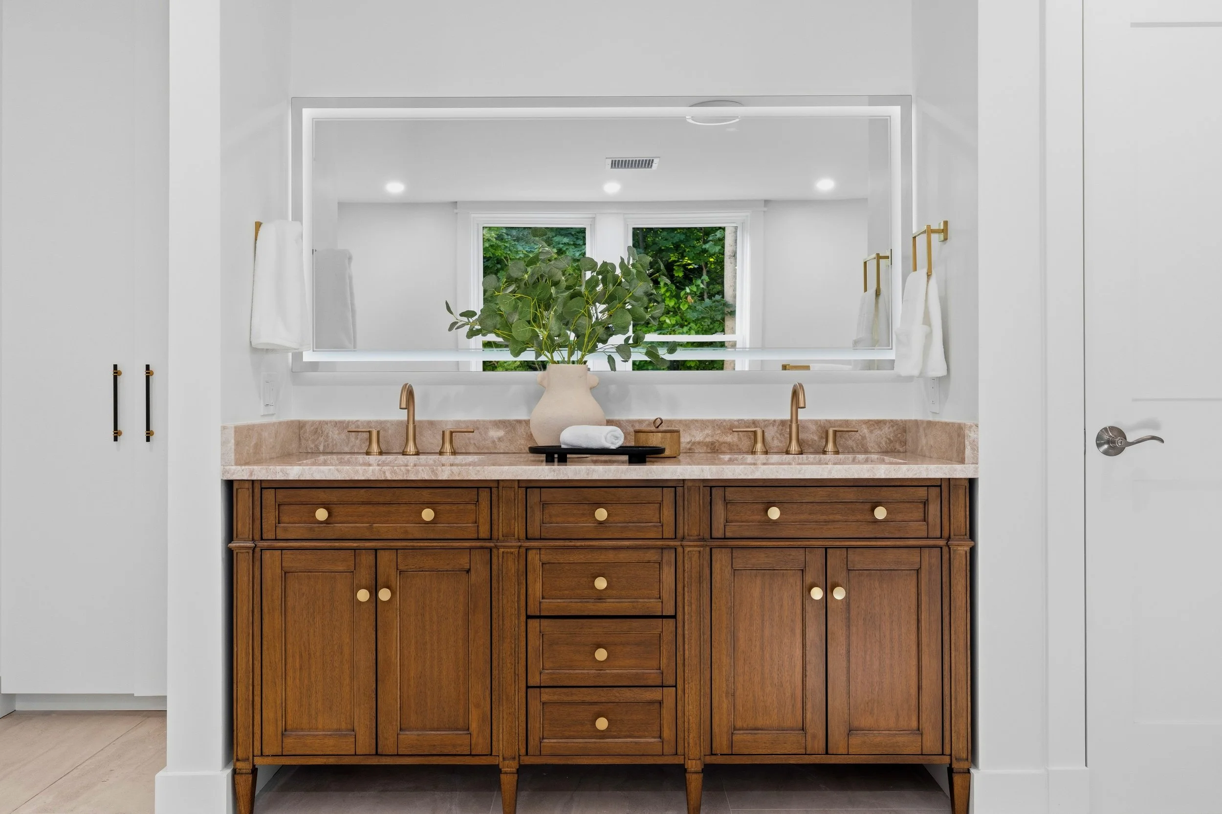 A bathroom vanity with a brown wood finish, multiple drawers, and gold knobs. A large mirror behind the vanity reflects a window with green foliage outside. On the vanity, there is a light-colored vase with green plants, a rolled white towel, and a s