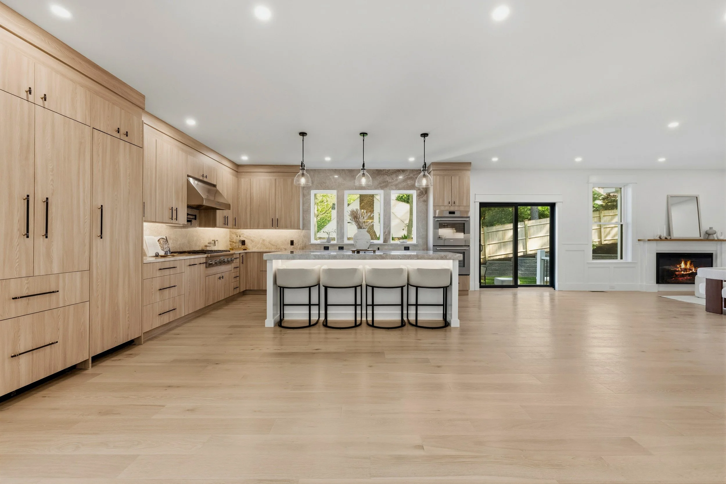 Spacious modern kitchen with light wood cabinetry, white island with seating, pendant lights, large windows, and sliding glass door leading outside.