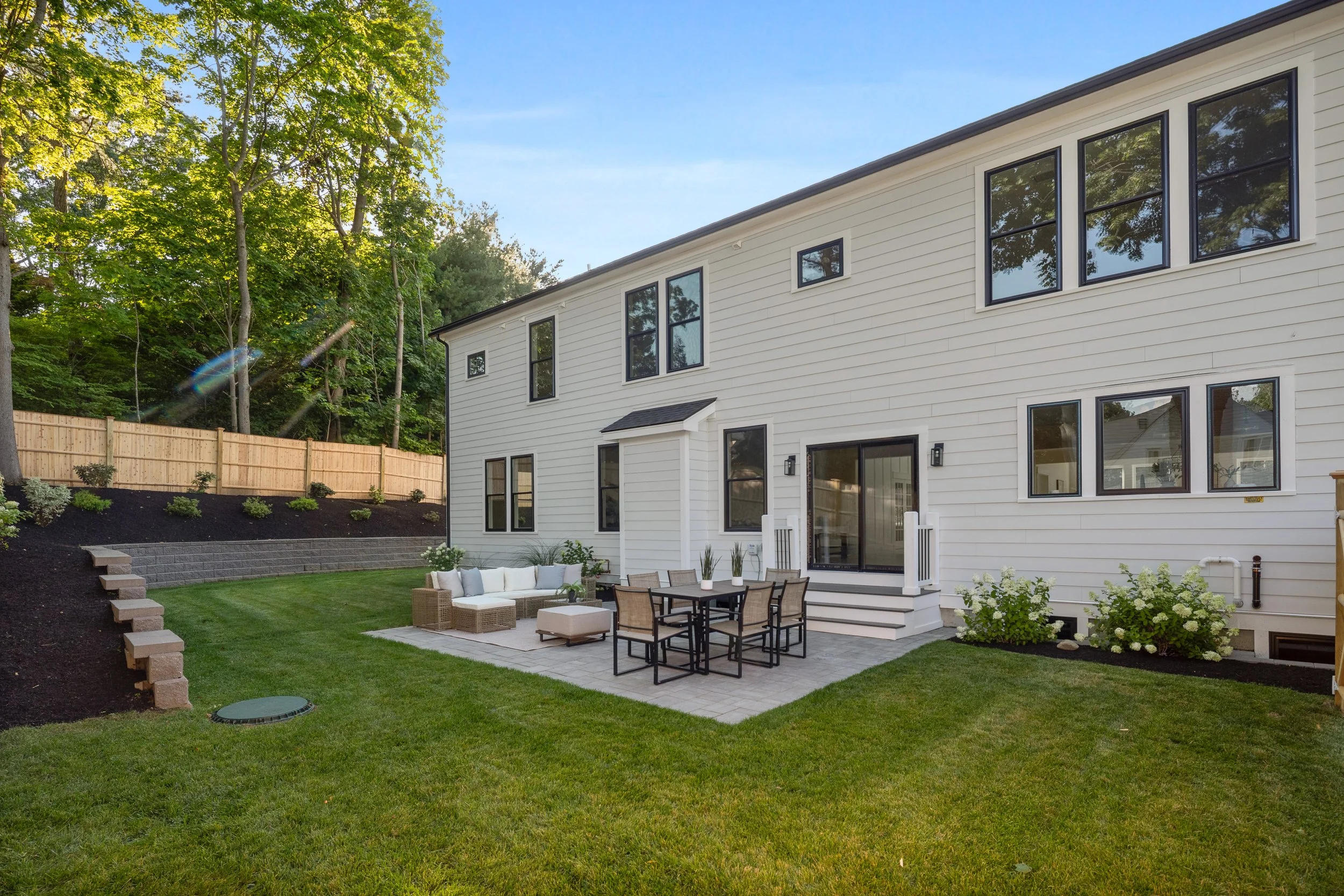 Backyard with outdoor furniture, green lawn, wooden fence, trees, and a white house with multiple windows.