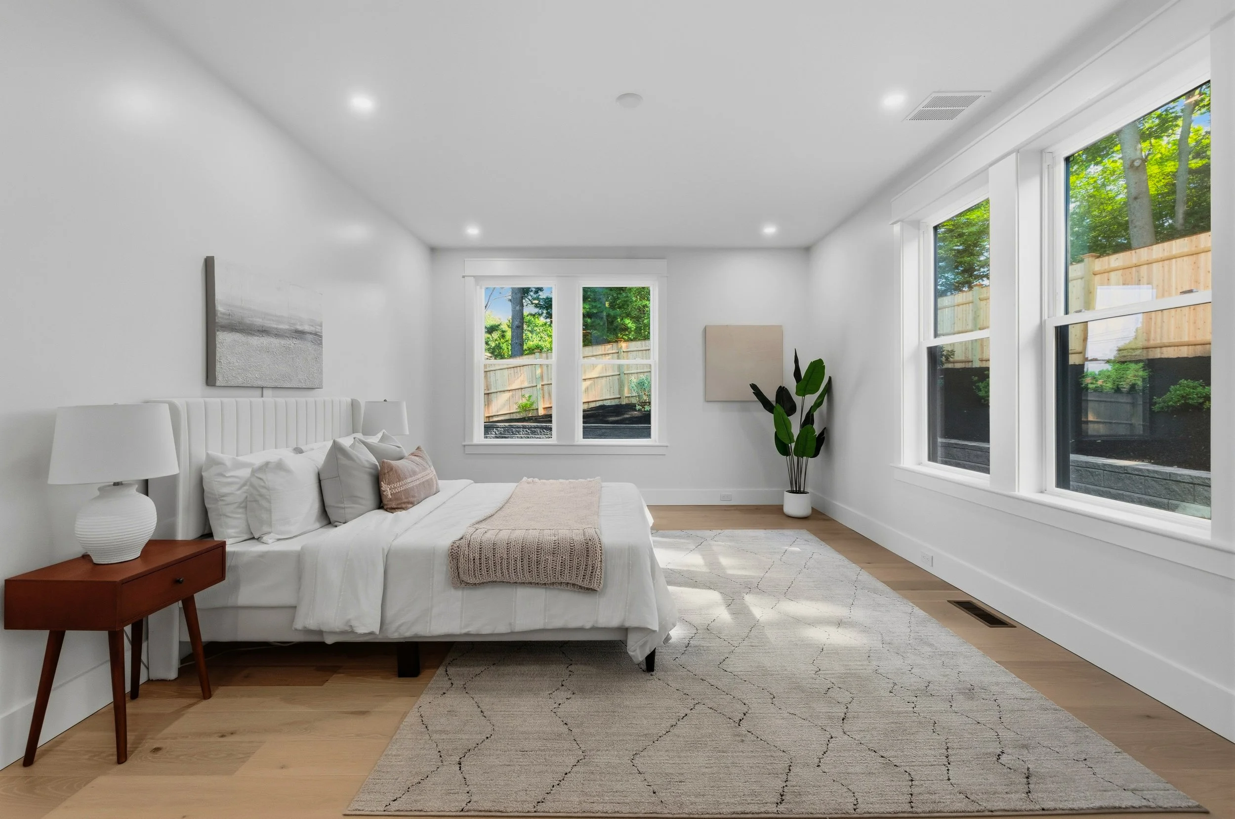 Bright modern bedroom with white walls, a white cushioned bed with pillows and a beige blanket, a wooden nightstand with a white ceramic lamp, large windows showing a fenced backyard with trees, and a potted plant near the window.